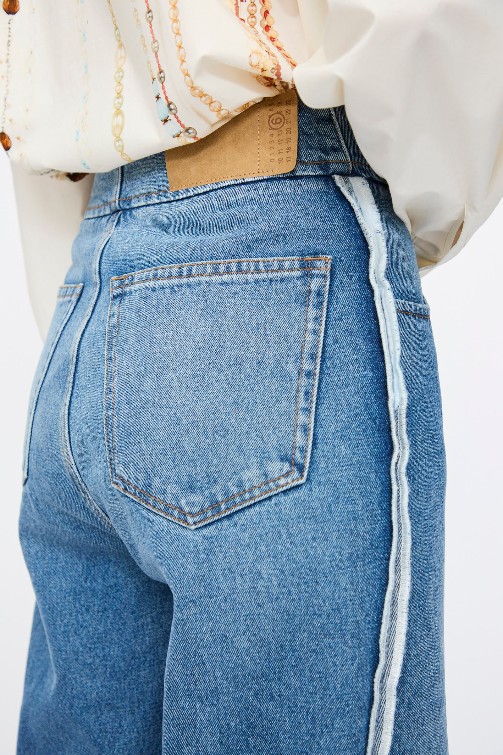 Close-up of a person wearing MM6 5 Pocket Pants in Medium Blue featuring a rear pocket and frayed side seam, paired with a white blouse decorated with beaded chains.