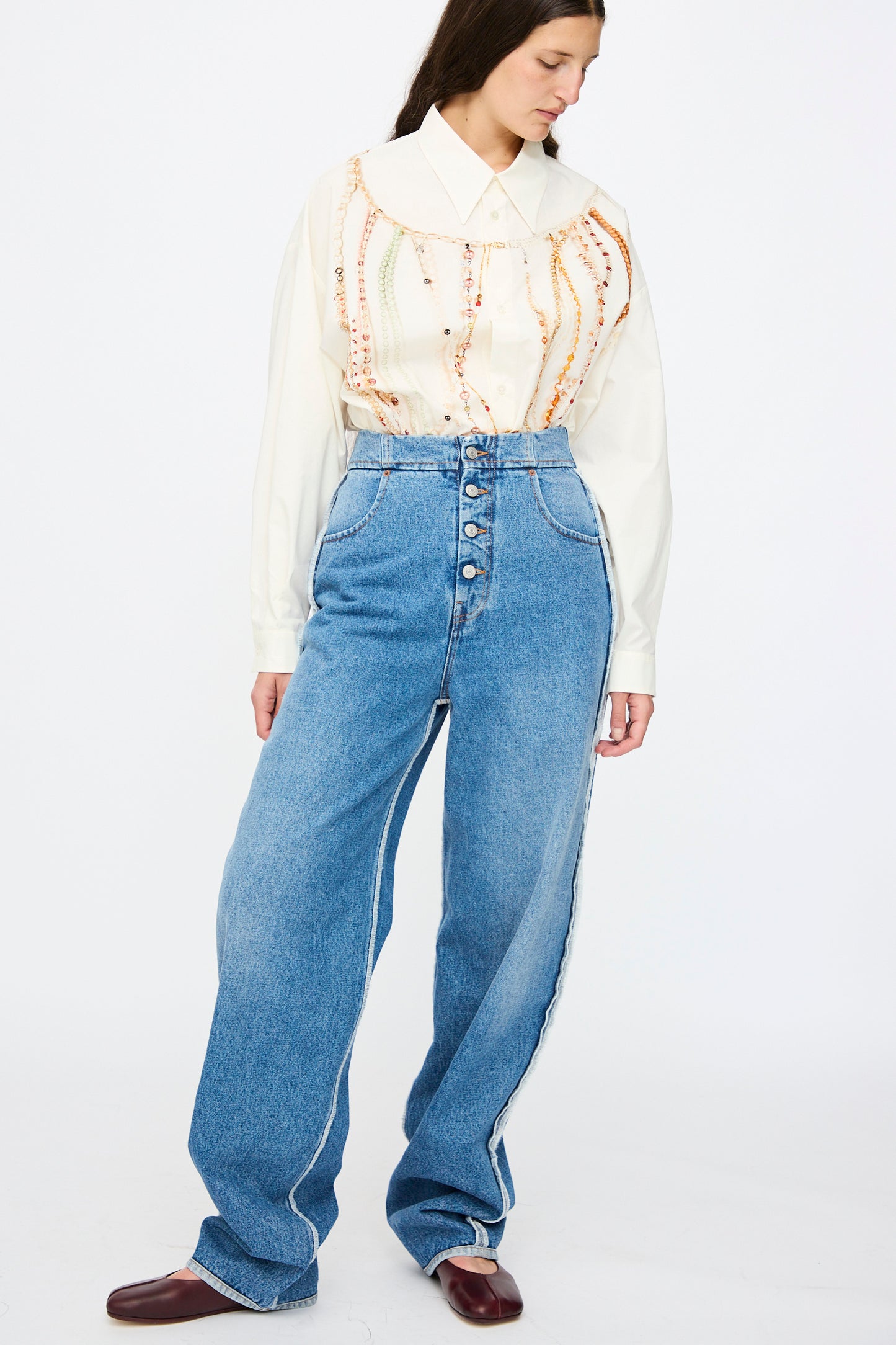 A woman stands against a plain background wearing a cream blouse, MM6 5 Pocket Pants in Medium Blue, and brown shoes.