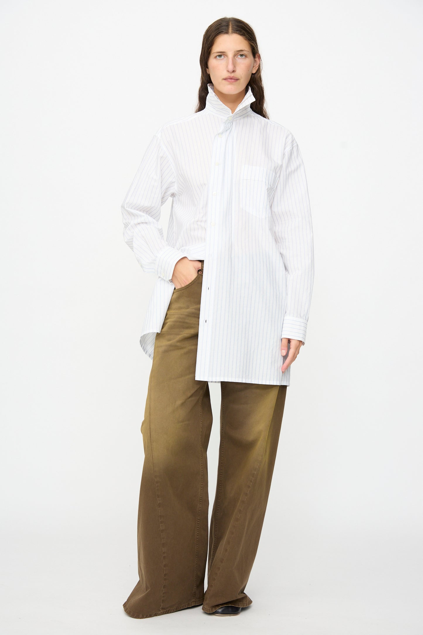 A person stands against a white background, wearing an oversized white pinstripe shirt with a high collar and relaxed fit, paired with MM6’s 5 Pockets Pants in Brown, with one hand in their pocket.