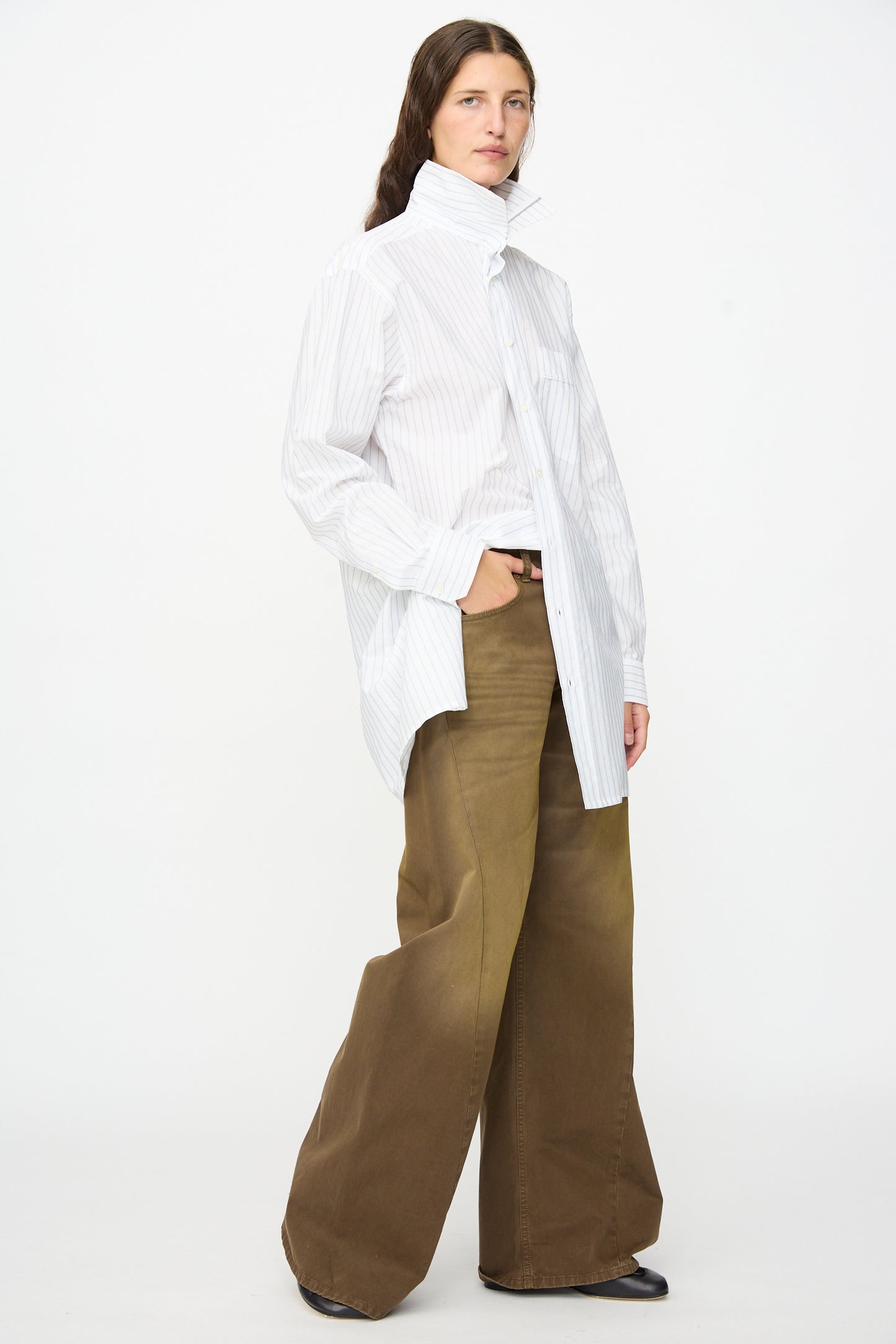A person stands against a plain background, hands in pockets, wearing an oversized white pinstripe shirt with a high collar and MM6′s 5 Pockets Pants in brown cotton twill denim, featuring a relaxed wide-leg fit.