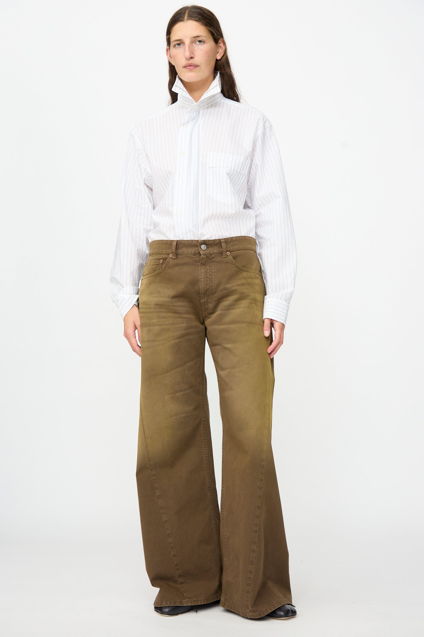 A person stands facing forward against a plain white background, wearing MM6′s 5 Pockets Pants in Brown paired with a white striped button-up shirt.
