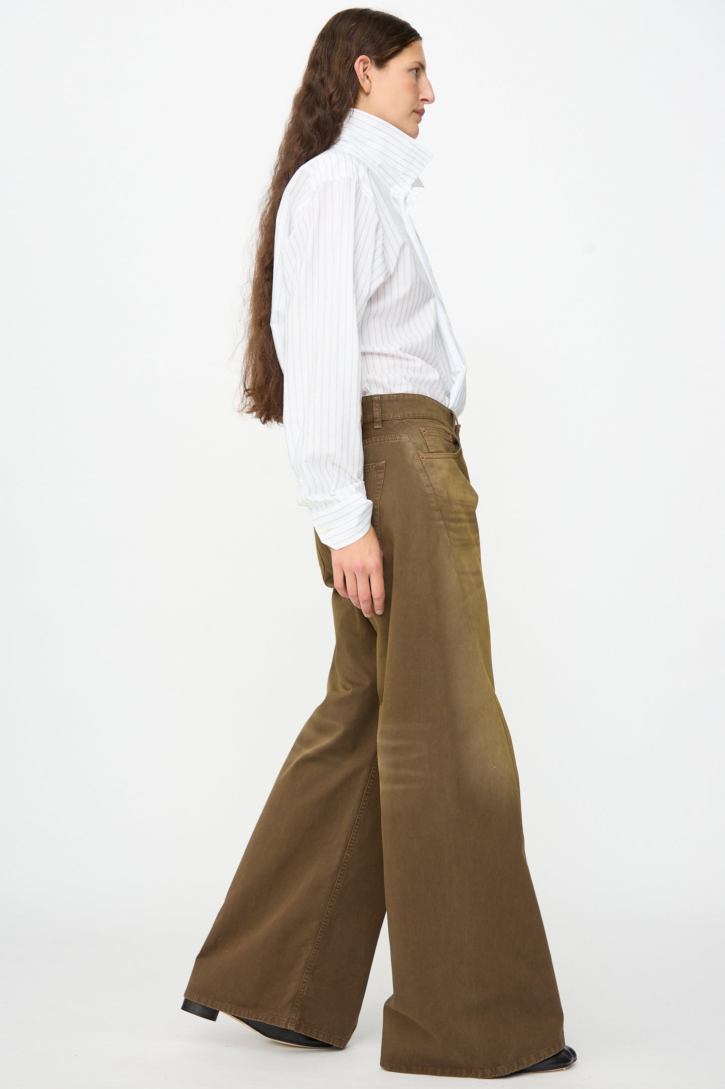 A person with long hair, wearing a white striped shirt and MM6 5 Pockets Pants in Brown, relaxed fit and wide leg in cotton twill denim, walks to the left against a plain white background.