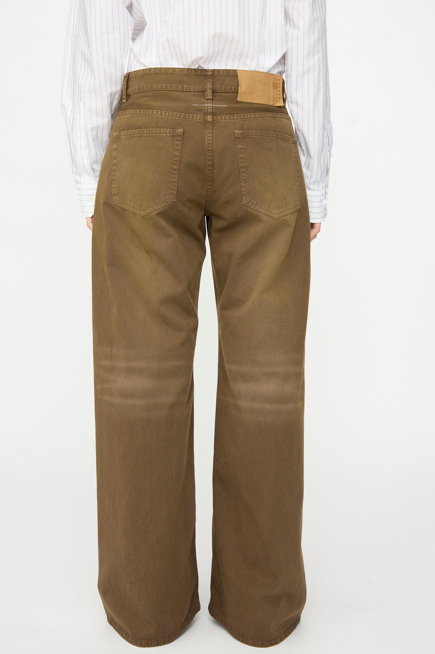 A person is shown from behind against a plain white background, wearing the MM6 5 Pockets Pants in Brown, featuring a relaxed fit and wide-leg silhouette in cotton twill denim, paired with a white striped shirt.