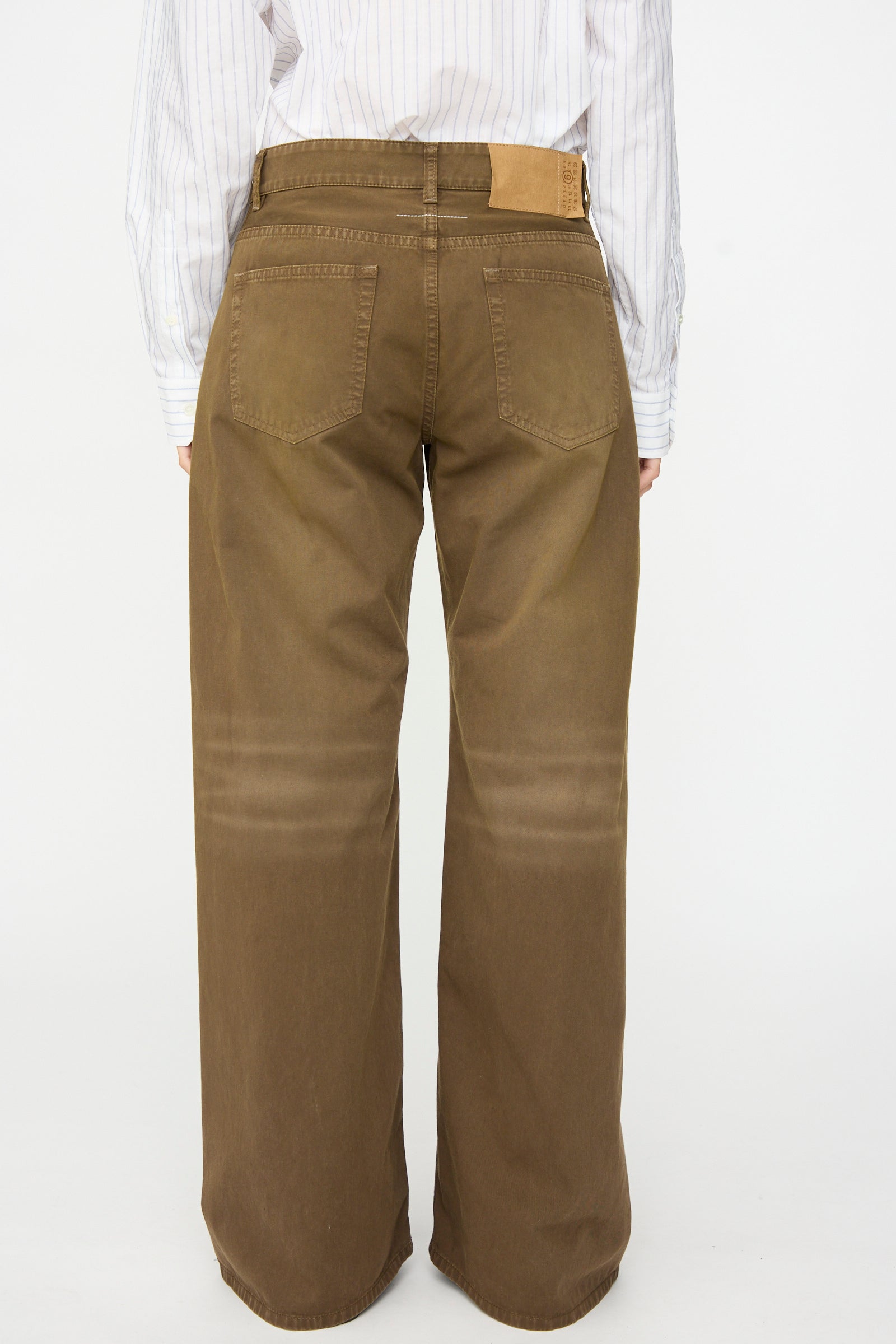 A person is shown from behind against a plain white background, wearing the MM6 5 Pockets Pants in Brown, featuring a relaxed fit and wide-leg silhouette in cotton twill denim, paired with a white striped shirt.