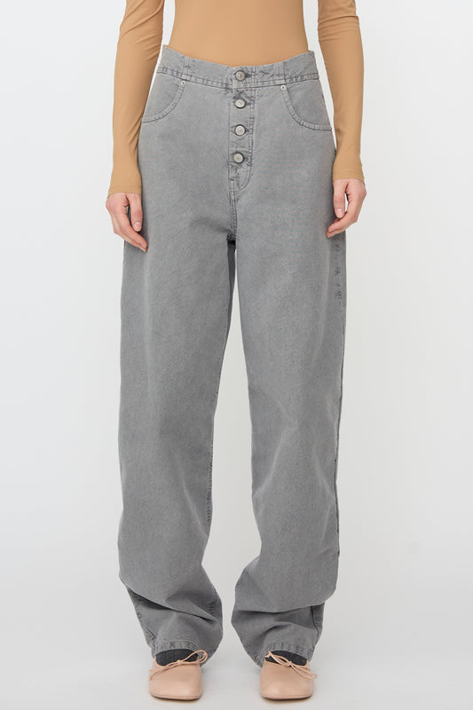 Someone wears MM6's 5 Pockets Pants in Grey, high-waisted with a relaxed fit and front buttons, paired with a beige long-sleeve top and light pink flats, standing against a plain background.