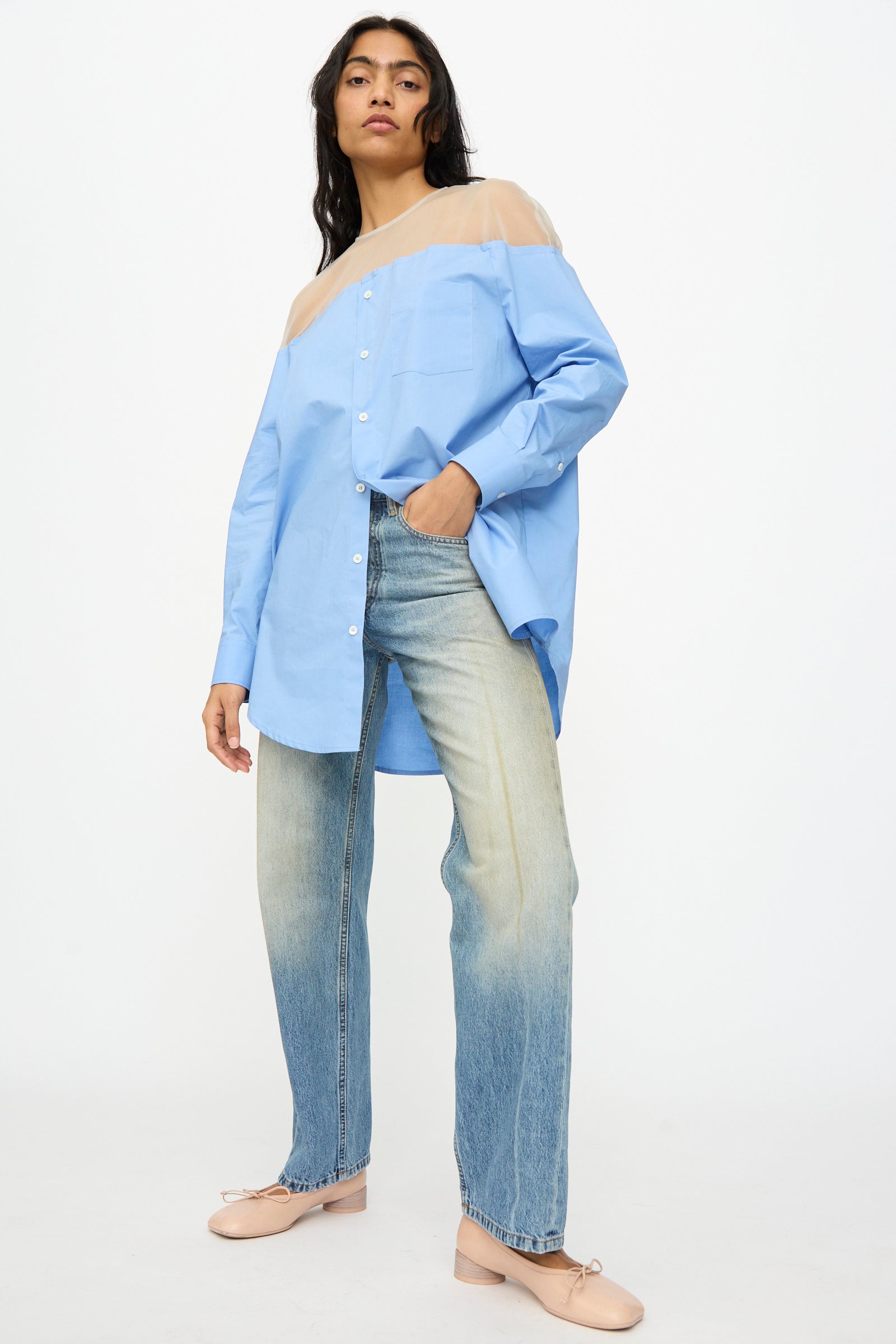 A woman stands against a plain background wearing MM6’s 5 Pockets Pants in Medium Blue, paired with a blue button-up shirt with sheer shoulders and pale pink flats.