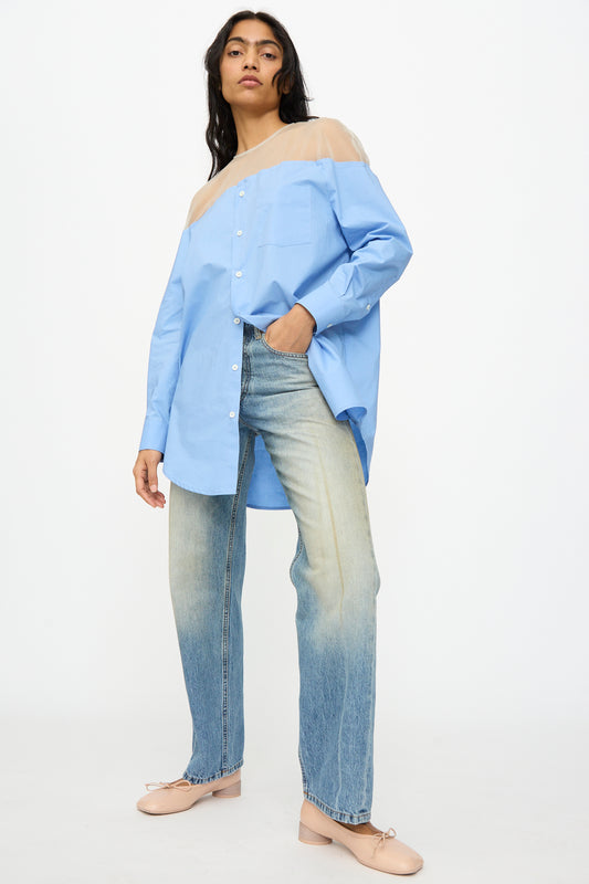 A woman stands against a plain background wearing MM6’s 5 Pockets Pants in Medium Blue, paired with a blue button-up shirt with sheer shoulders and pale pink flats.