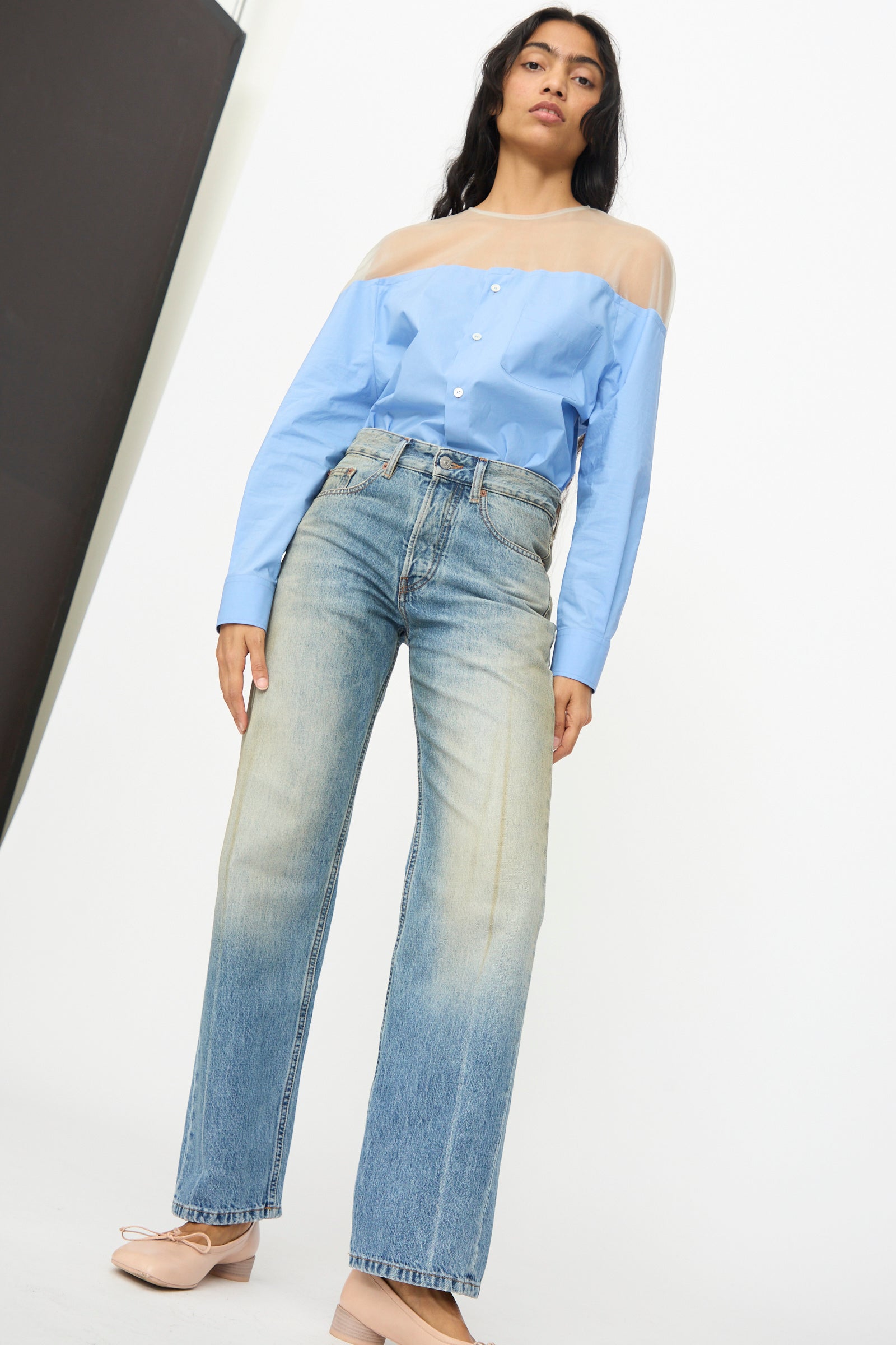 A person stands against a white background wearing a blue long-sleeve top with sheer shoulders, MM6 5 Pockets Pants in Medium Blue, and light pink shoes.