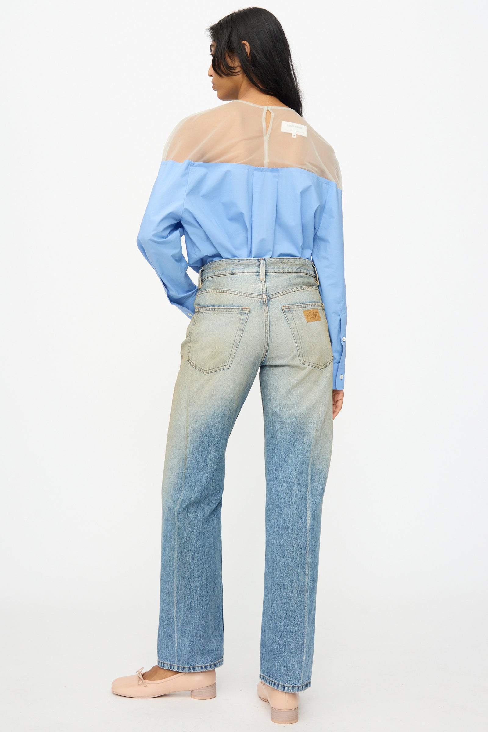 A person stands facing away, wearing a blue shirt with a sheer beige yoke, MM6 5 Pockets Pants in Medium Blue, and light pink shoes.