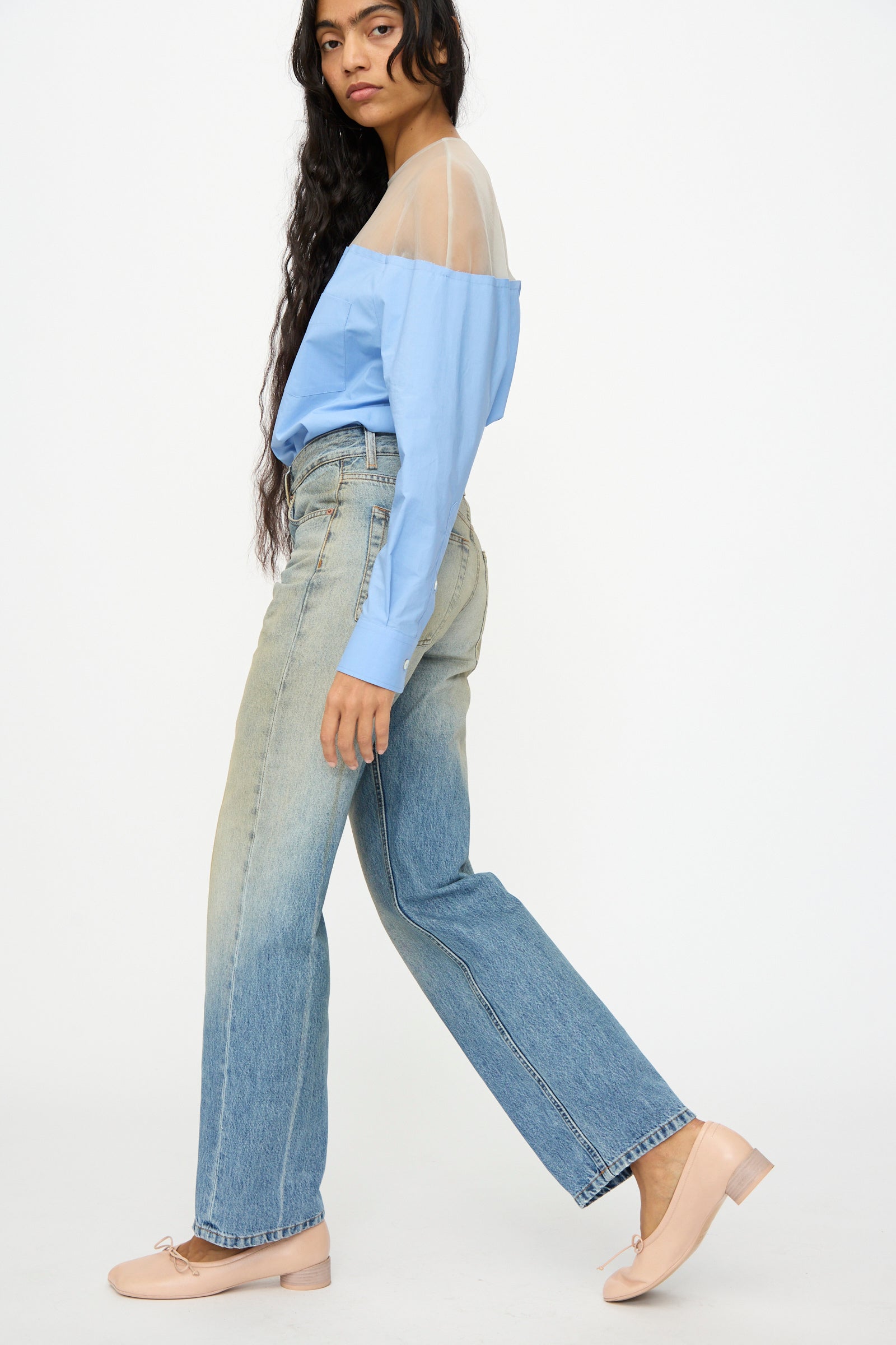 A woman stands sideways against a plain white background, wearing MM6 5 Pockets Pants in Medium Blue, a blue off-the-shoulder sheer top, and beige flats.