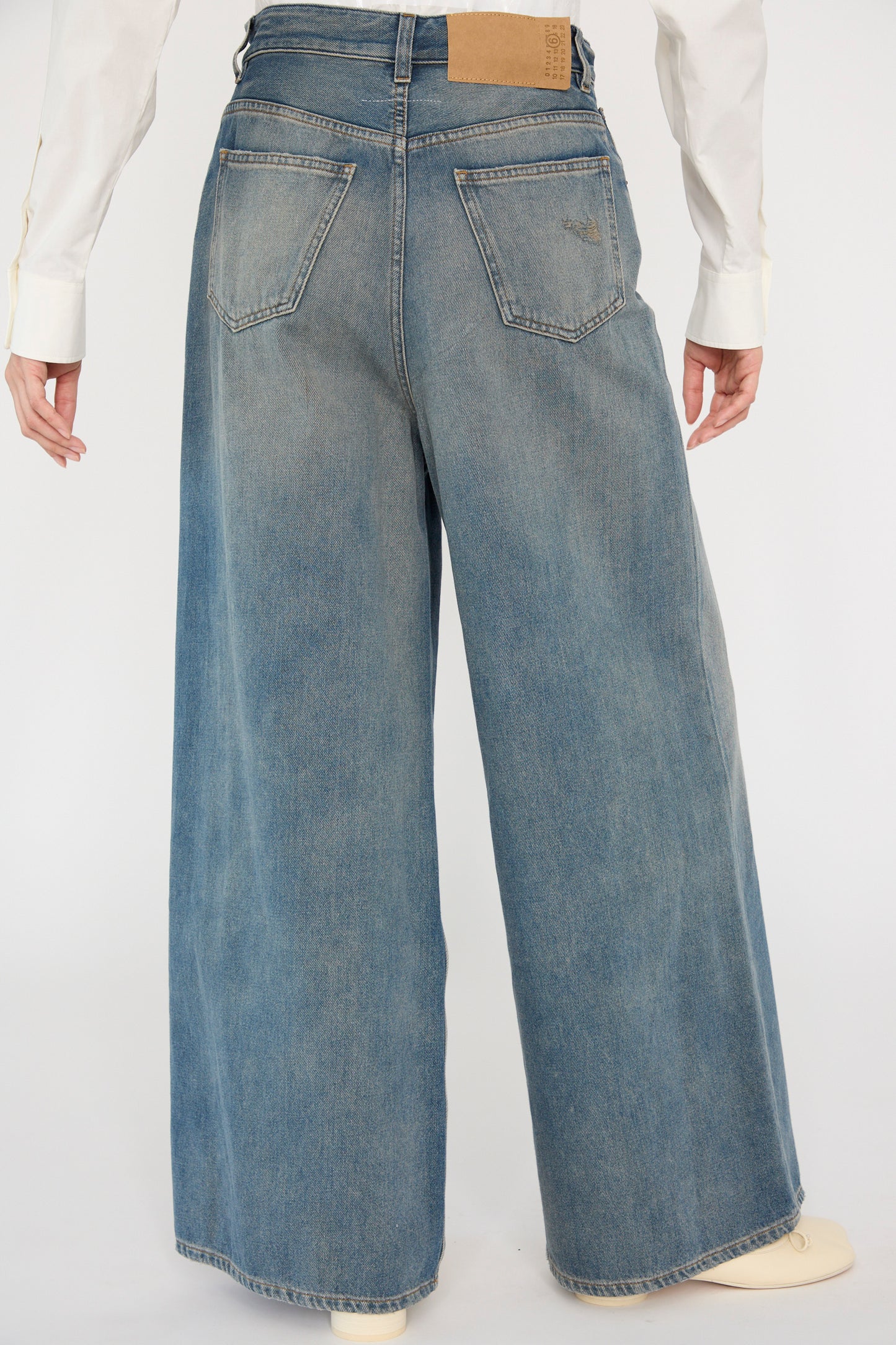 A person in MM6's 5 Pocket Pant in Grey Blue and a white shirt is seen from behind. These high-rise, wide-leg jeans with back pockets and a leather patch on the waistband offer a stylish blend of modern and classic design.