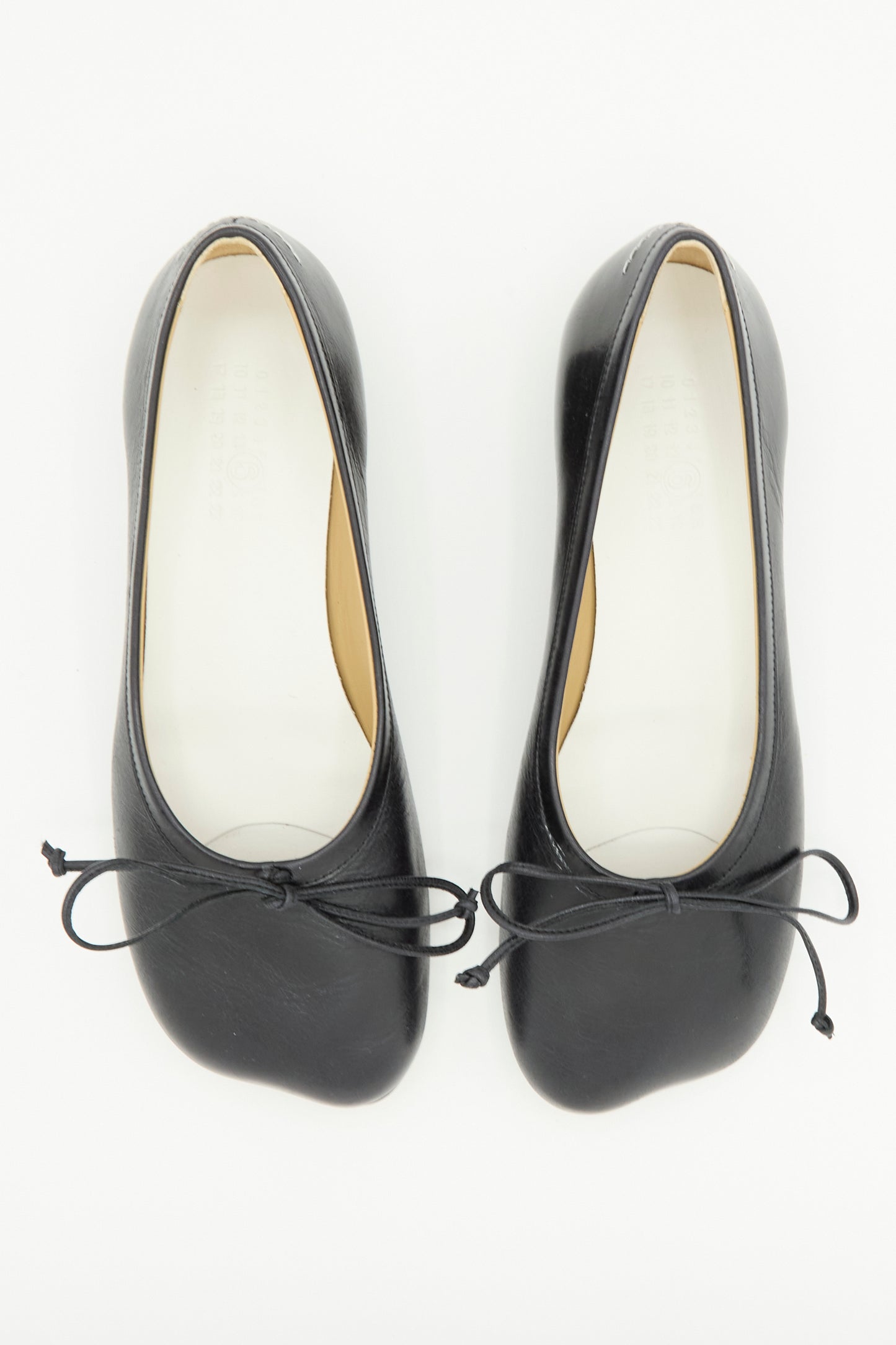 A pair of MM6 Ballet Shoes in Black, featuring rounded toes and delicate front bows, expertly crafted from supple calf leather and photographed from above against a white background.