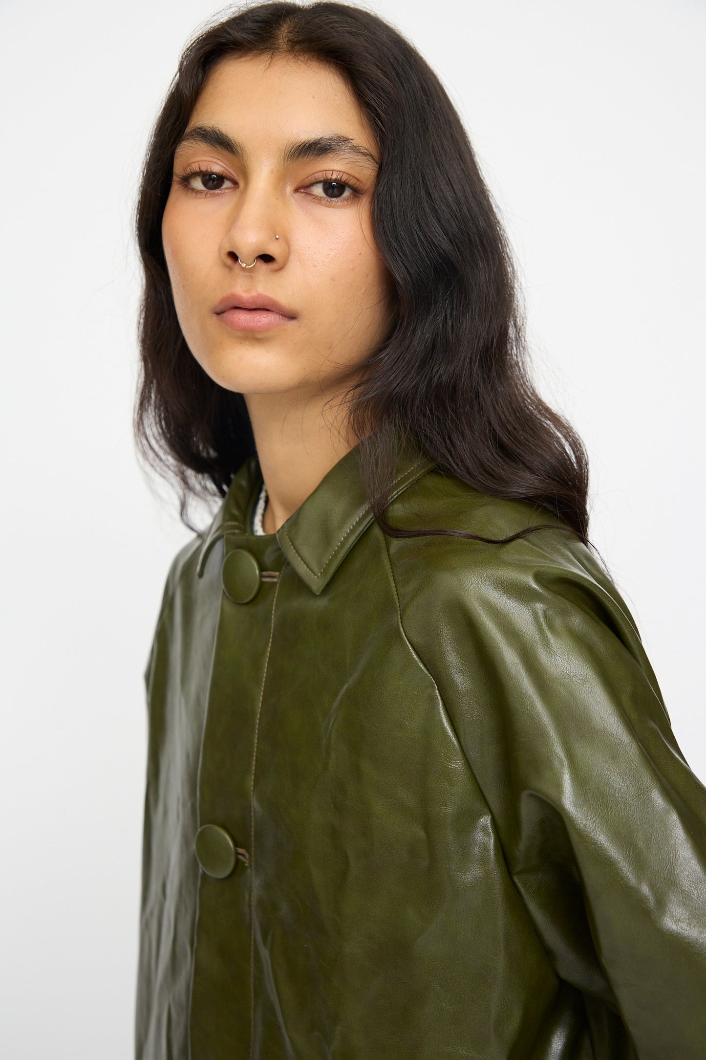 Wearing the MM6 Coat in Forest Green, a person with long dark hair and a nose ring looks directly at the camera against a plain white background.