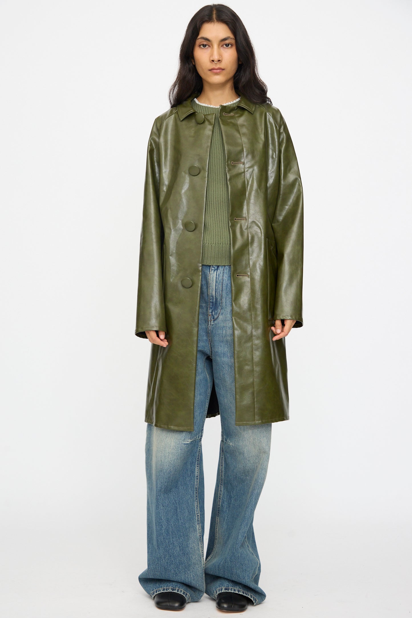 A person models the MM6 Coat in Forest Green over a green sweater and wide-leg blue jeans, standing against a plain white background.