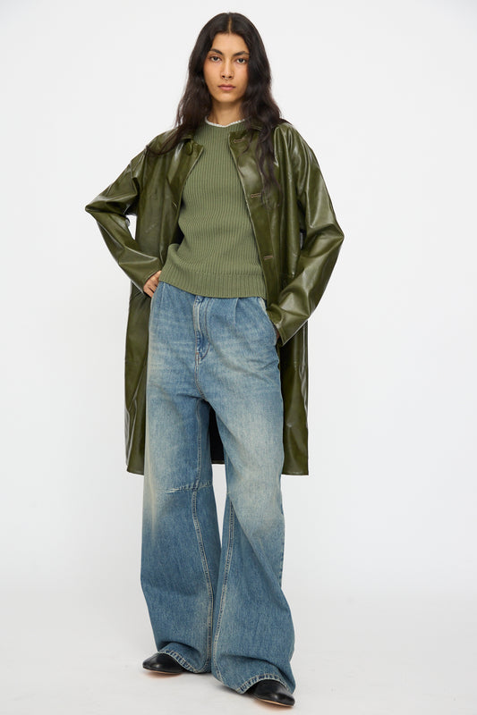 A person faces forward wearing the MM6 Coat in Forest Green, paired with a ribbed green sweater, wide-leg faded blue jeans, and black shoes.