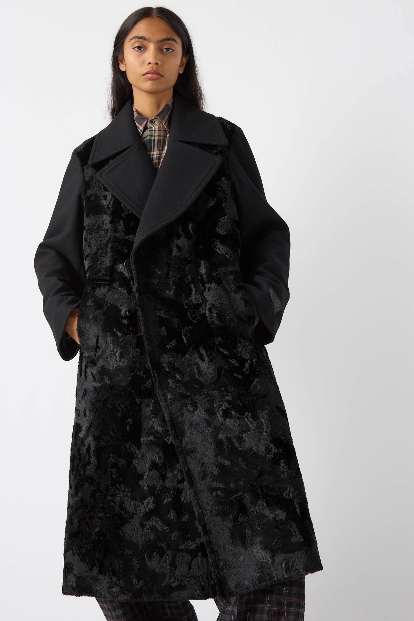 A person stands wearing the MM6 Coat in Black, a long, textured wool blend coat with broad lapels over a plaid shirt. The coat has a fur-like appearance and extends below the knees.