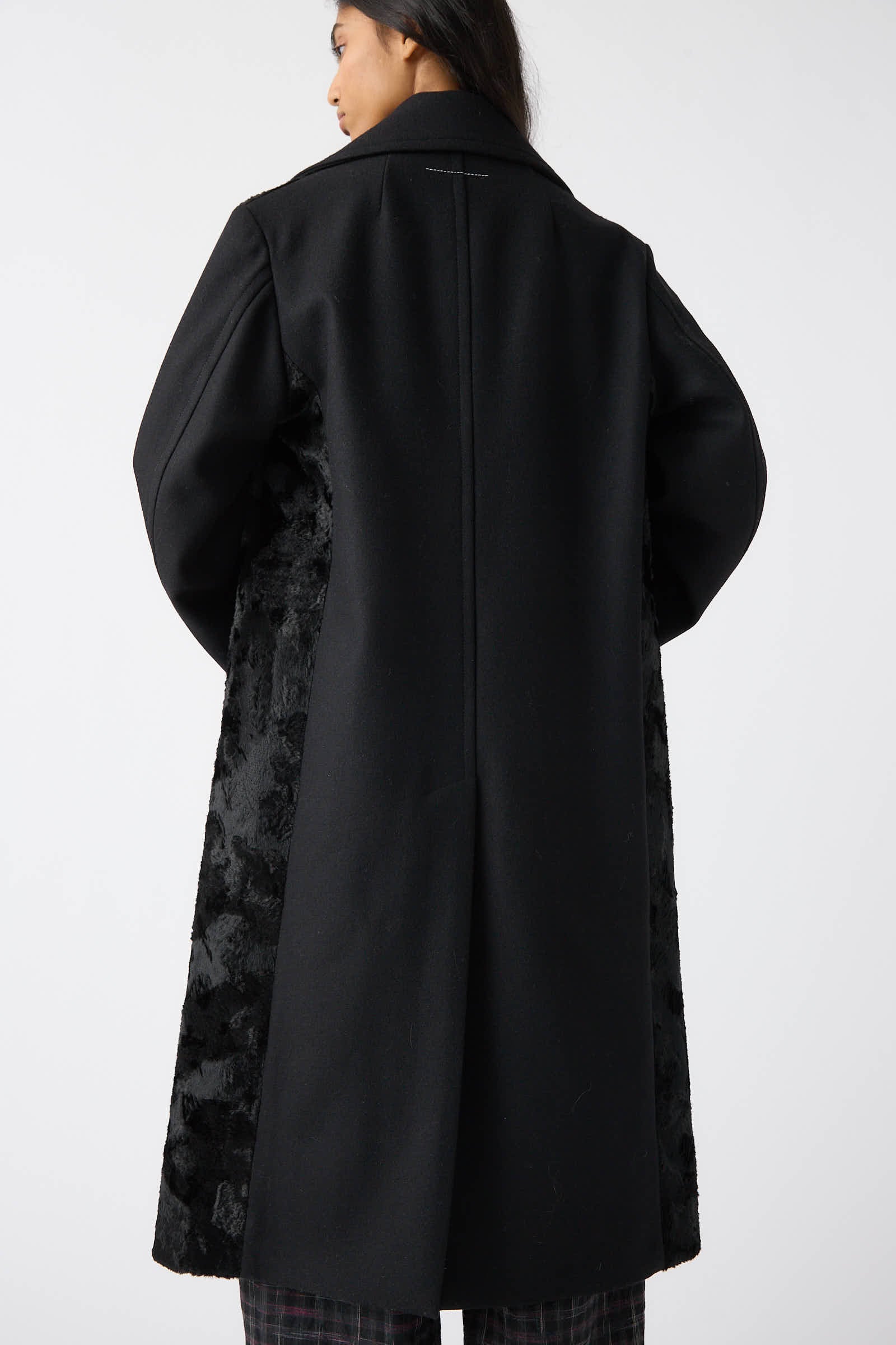 A person wearing the MM6 Coat in Black with broad lapels, photographed from behind. The coat features textured side panels, and the individual has long hair.