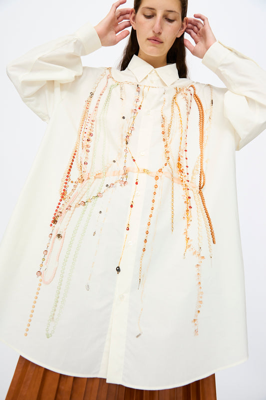 A person wearing the MM6 Dress in Pearls Print, an oversized white shirt featuring a pearls-inspired design, paired with a brown pleated skirt.