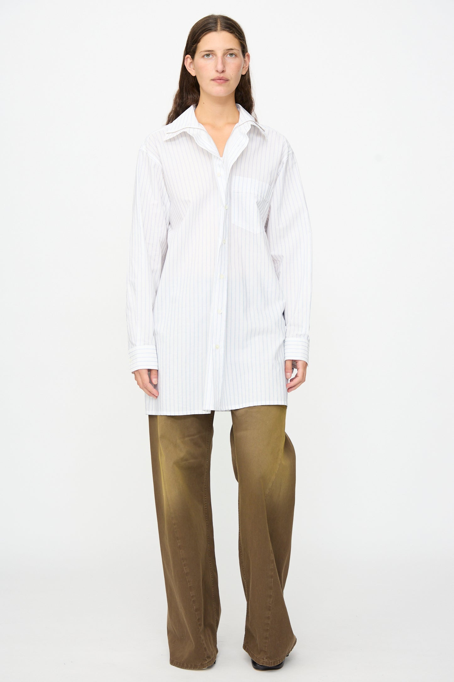 A person stands facing forward against a plain white background, wearing the MM6 Long-Sleeved Shirt in White and Blue—crafted in Italy from 100% cotton with pinstripes—and paired with wide-leg brown pants.