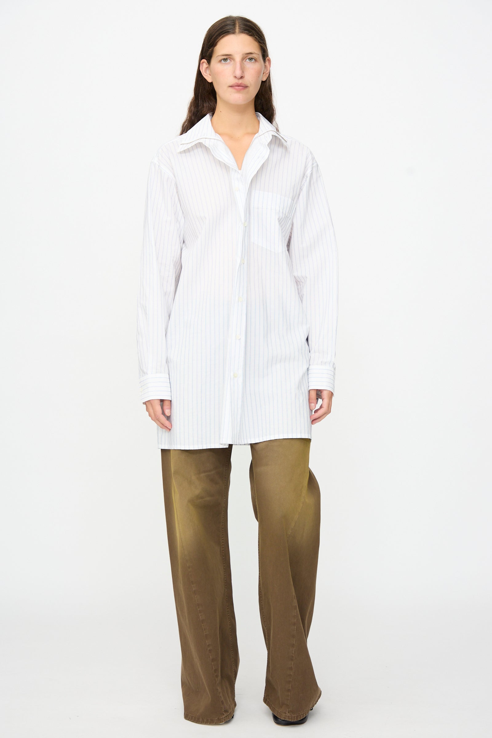 A person stands facing forward against a plain white background, wearing the MM6 Long-Sleeved Shirt in White and Blue—crafted in Italy from 100% cotton with pinstripes—and paired with wide-leg brown pants.