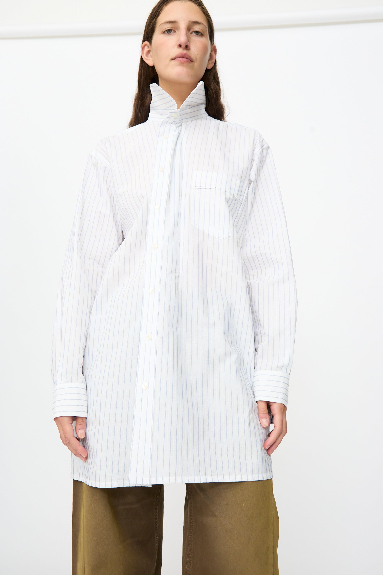 A person wearing the MM6 Long-Sleeved Shirt in White and Blue—an oversized, 100% cotton piece with a high collar and pinstripe design—paired with brown wide-leg pants, stands against a plain white background.