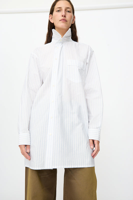 A person wearing the MM6 Long-Sleeved Shirt in White and Blue—an oversized, 100% cotton piece with a high collar and pinstripe design—paired with brown wide-leg pants, stands against a plain white background.