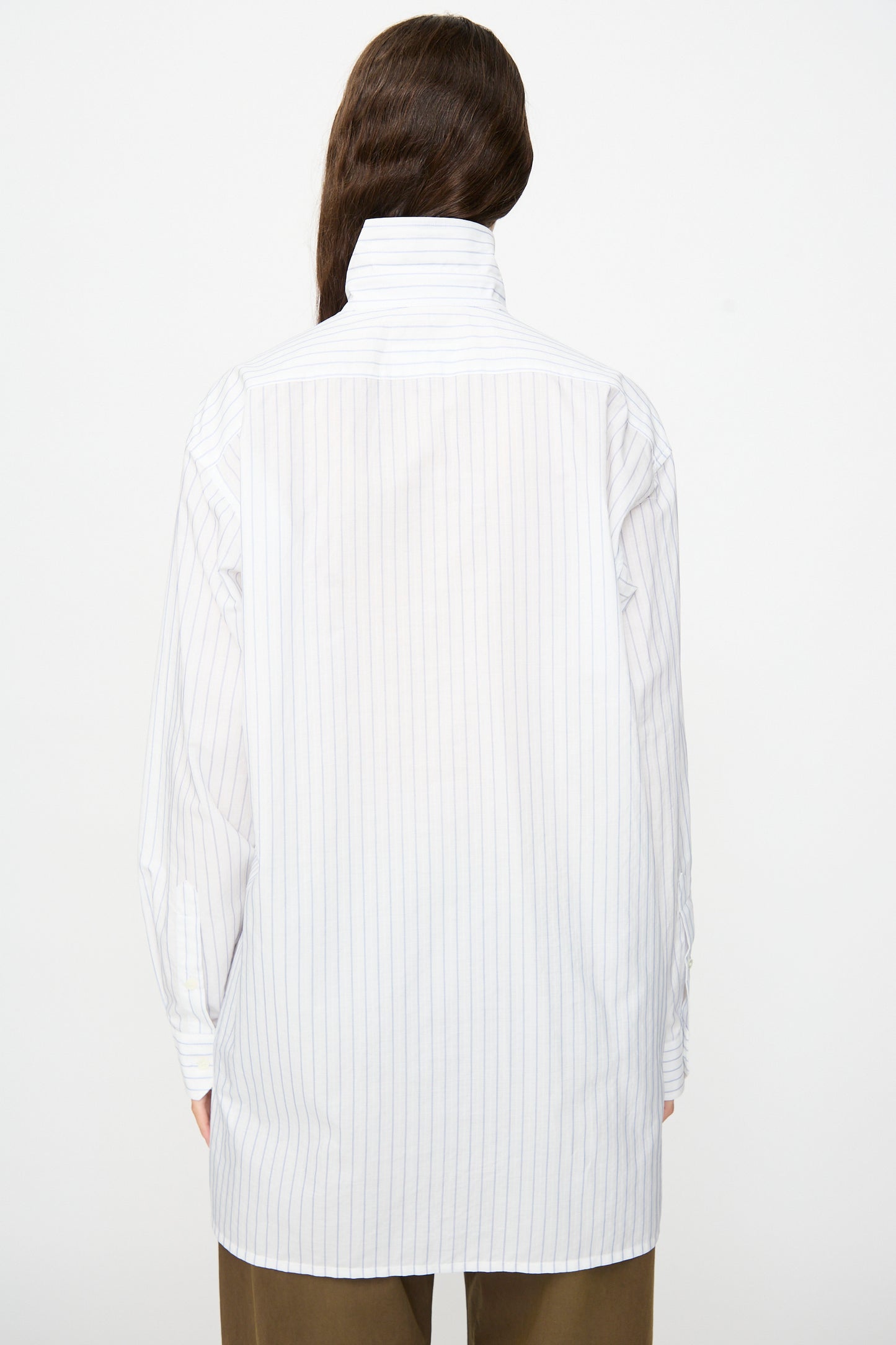 A person stands facing away, wearing the MM6 Long-Sleeved Shirt in White and Blue, featuring thin vertical pinstripes and a 100% cotton construction with the collar turned up. They are also wearing brown pants, with long wavy hair visible against a plain white background.