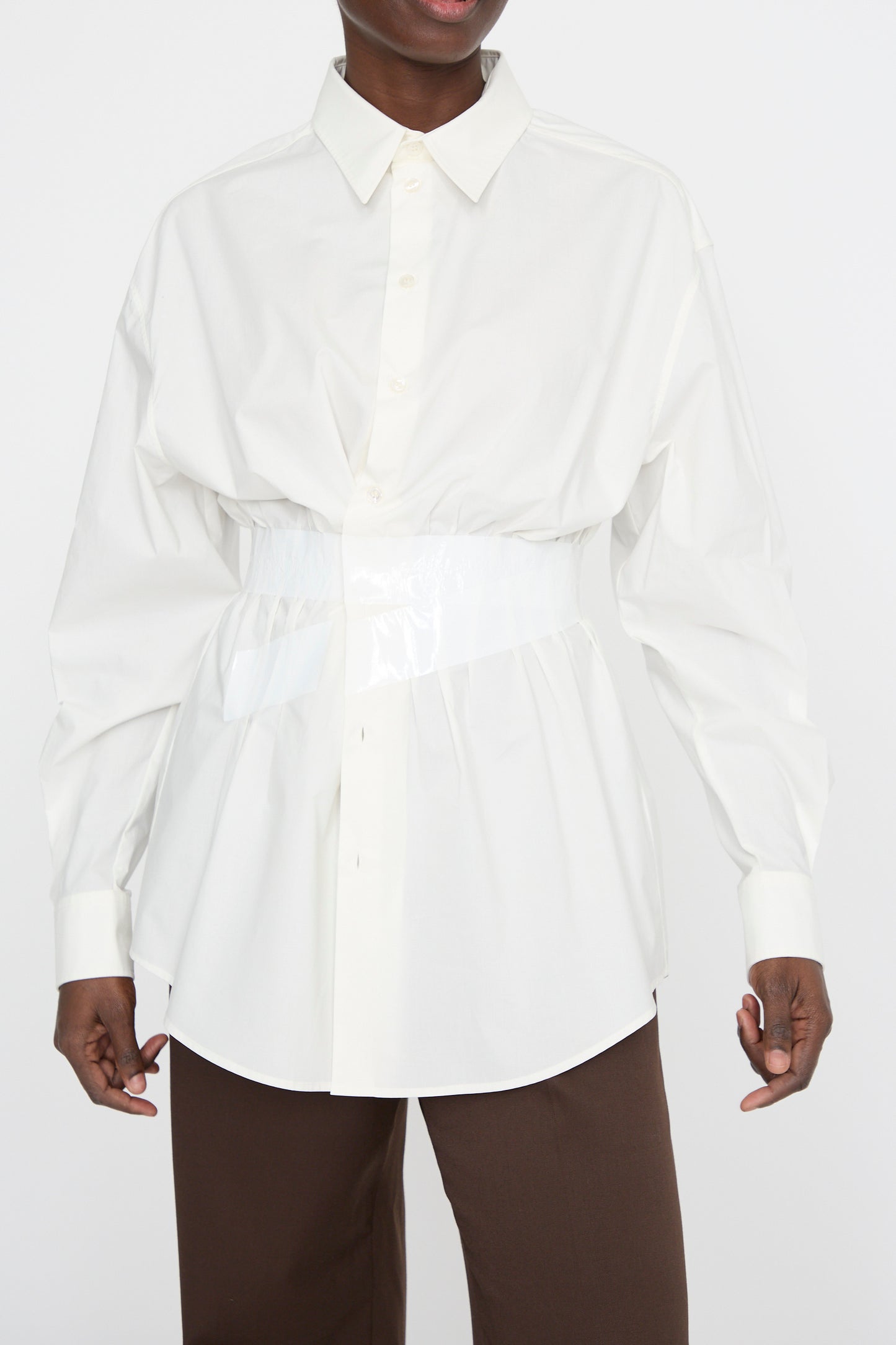 A person wears MM6's Long Sleeve Shirt in Off White, a relaxed-fit button-up with a glossy cinched waist, paired with brown pants against a plain background.