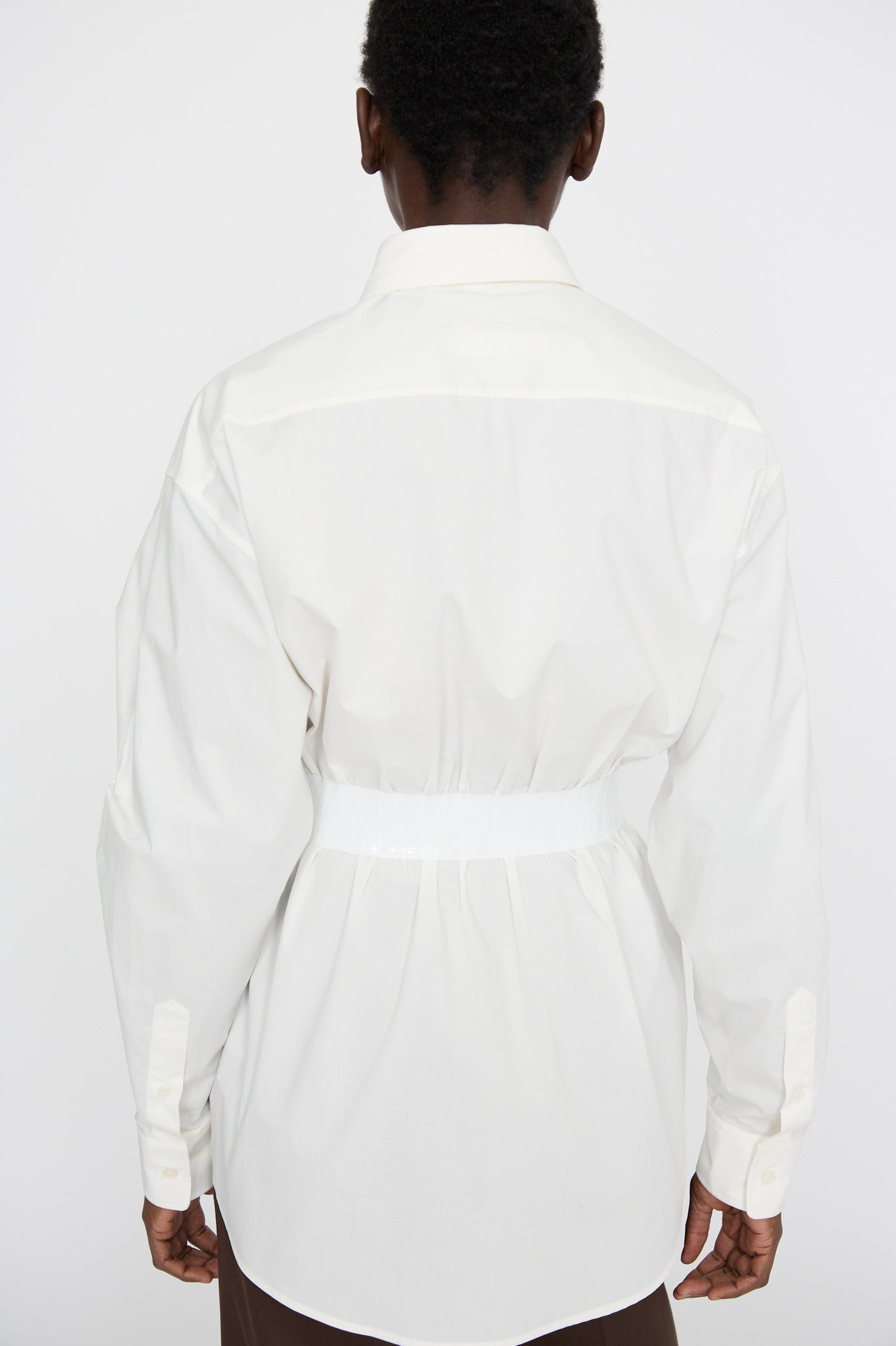 An individual is spotted in a MM6 Long Sleeve Shirt in Off White, characterized by its relaxed fit and crafted from cotton poplin. The shirt includes an elegant back belt detail, offering a refined yet casual rear view.