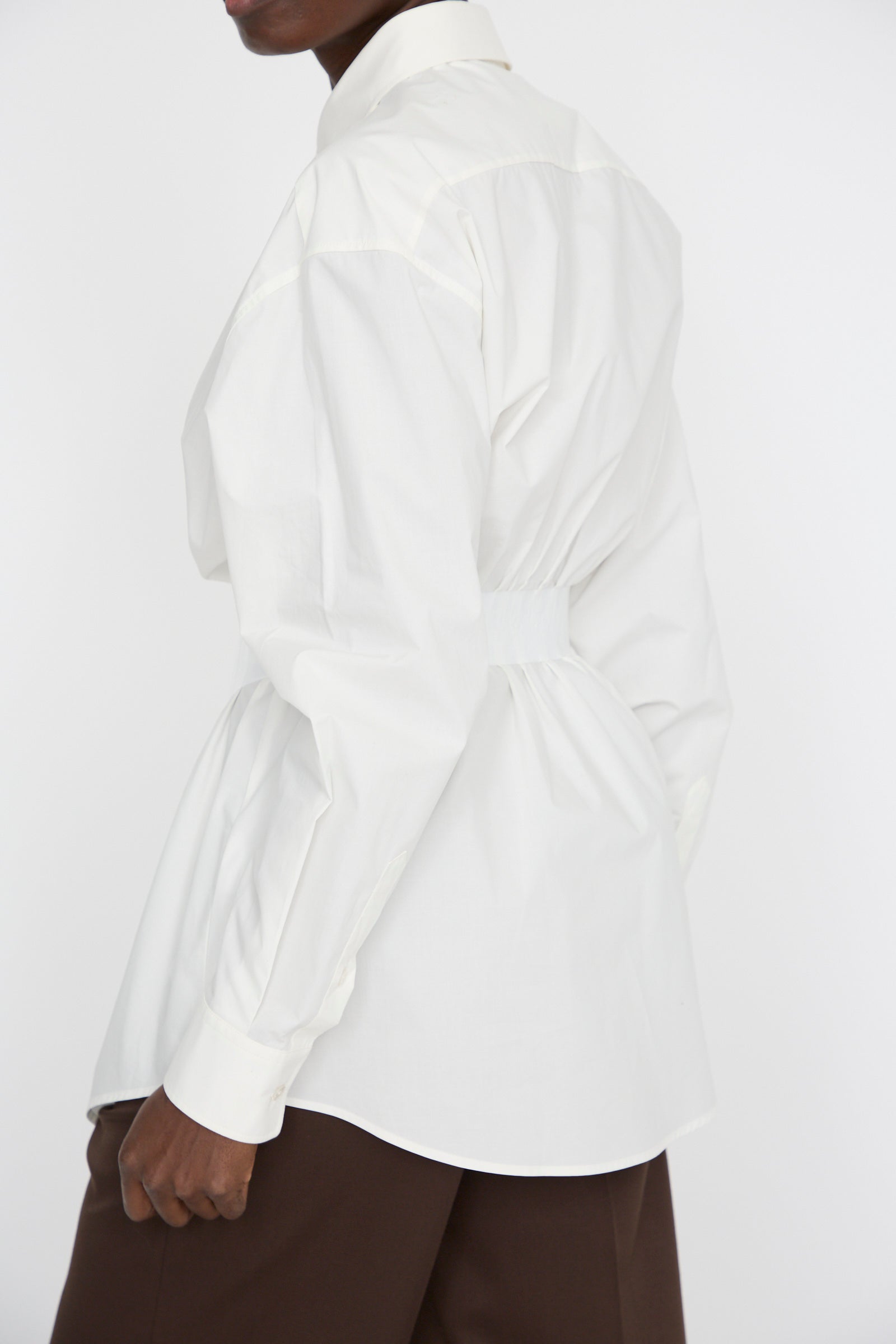 Effortlessly dressed in an MM6 Long Sleeve Shirt in Off White, featuring a relaxed fit and cinched waist, a person pairs it with brown pants. They stand sideways against a light background.