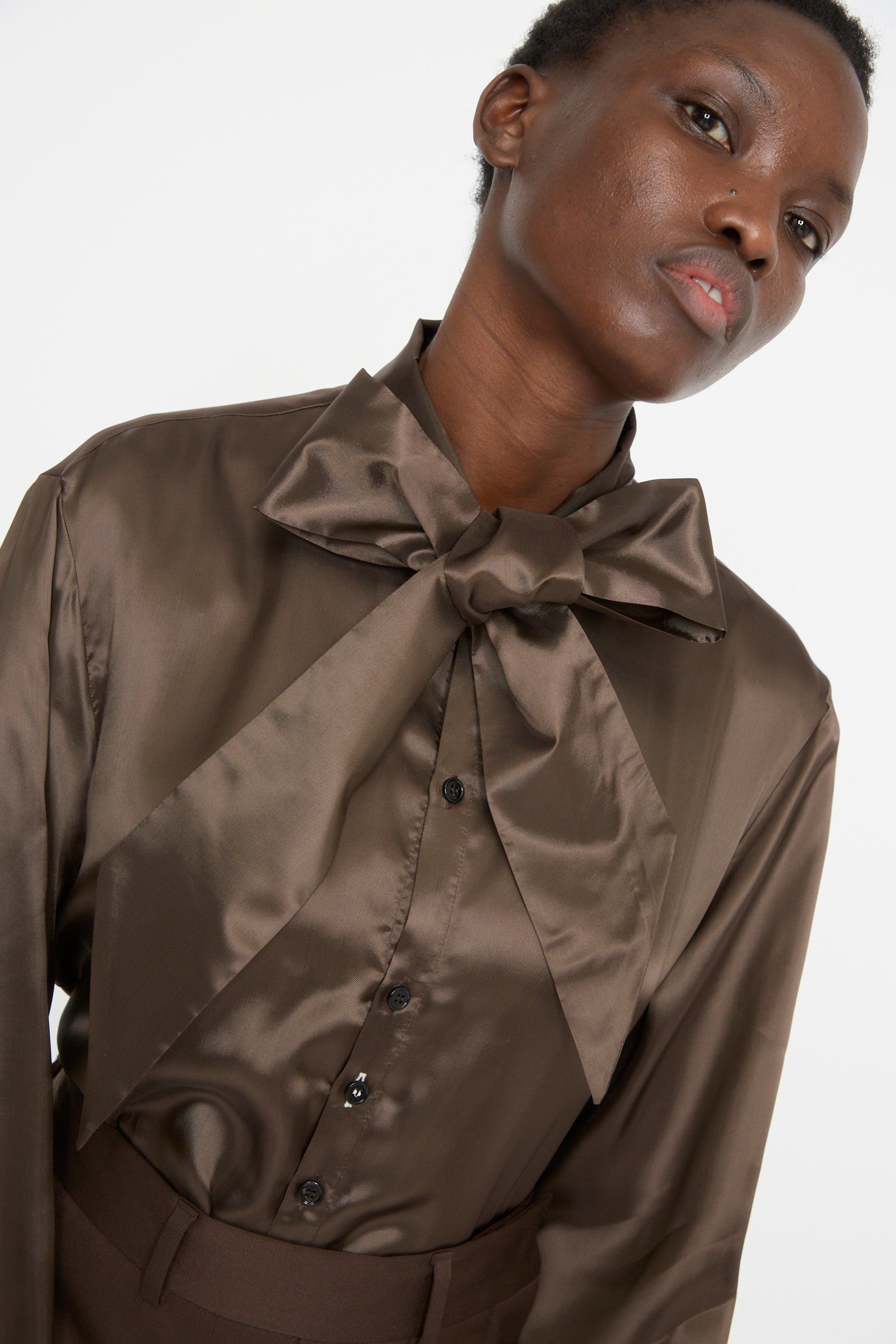 A person stands against a white background wearing the MM6 Long Sleeved Shirt in Dark Brown. Made of viscose satin with a bow tie neck, the shirt drapes elegantly, adding sophistication to the look.