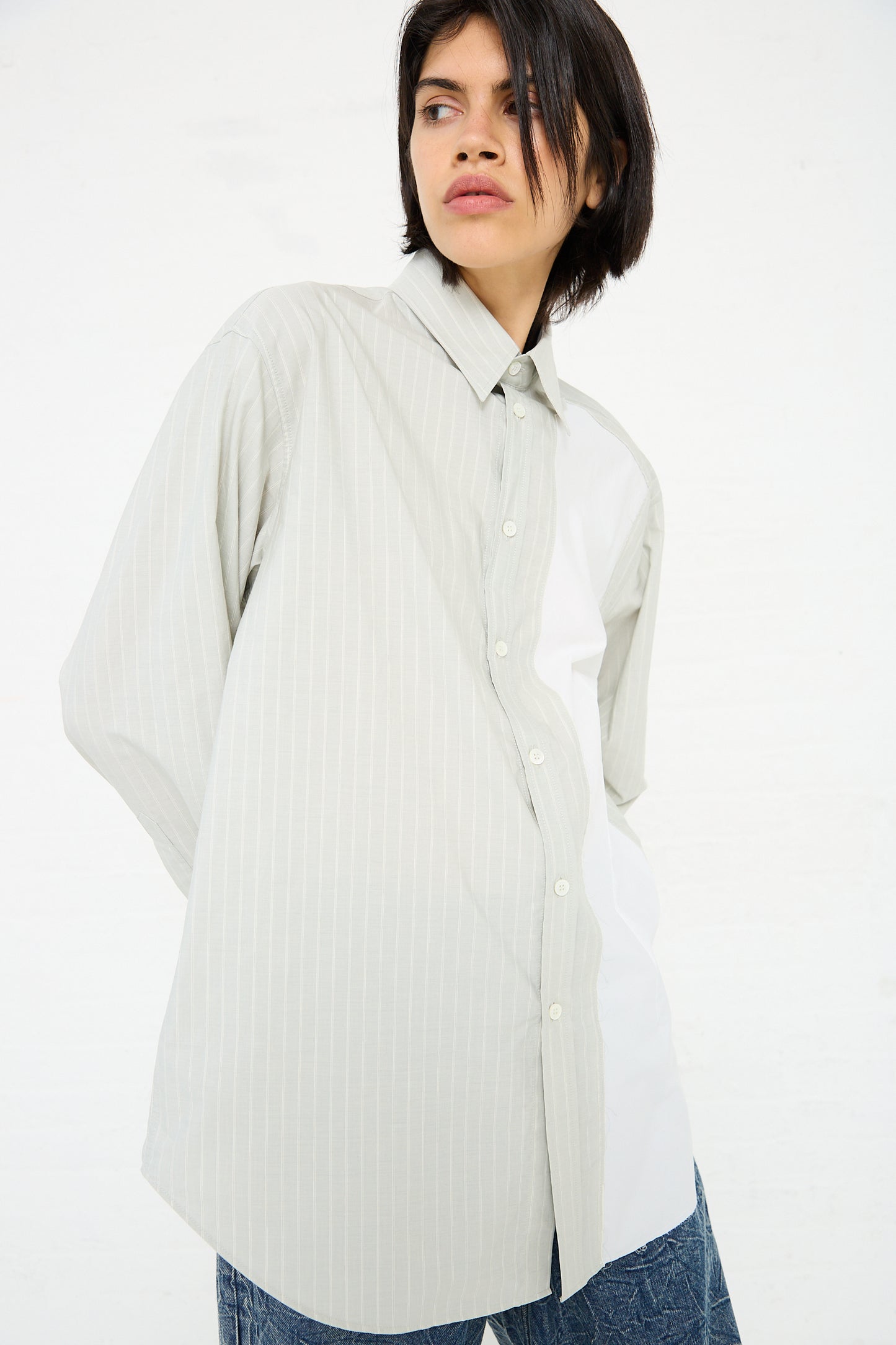 Person wearing a loose-fitting, light-colored, 100% MM6 Long Sleeved Shirt in Iguana and blue jeans stands against a plain white background looking to the side.