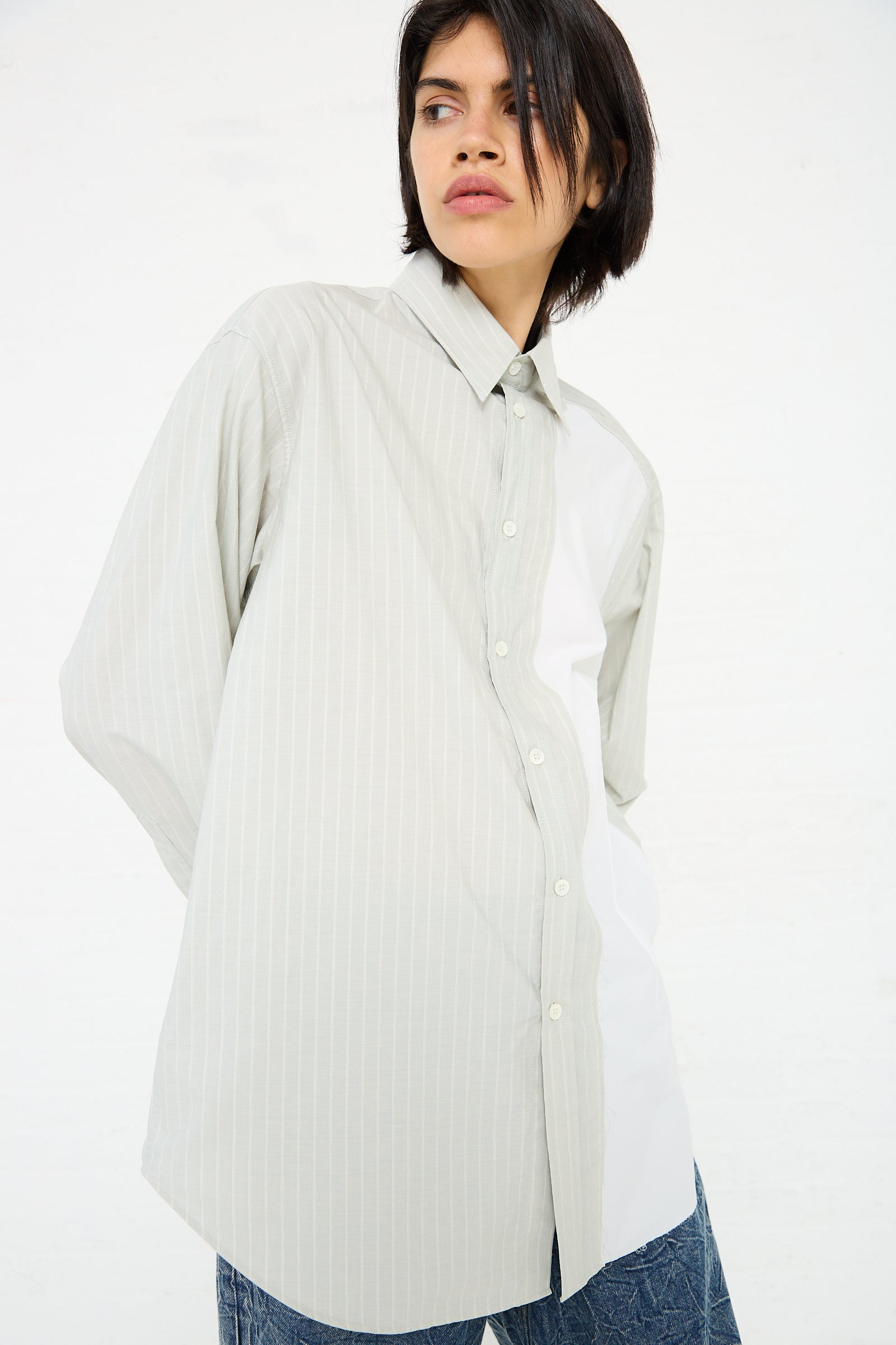 Person wearing a loose-fitting, light-colored, 100% MM6 Long Sleeved Shirt in Iguana and blue jeans stands against a plain white background looking to the side.