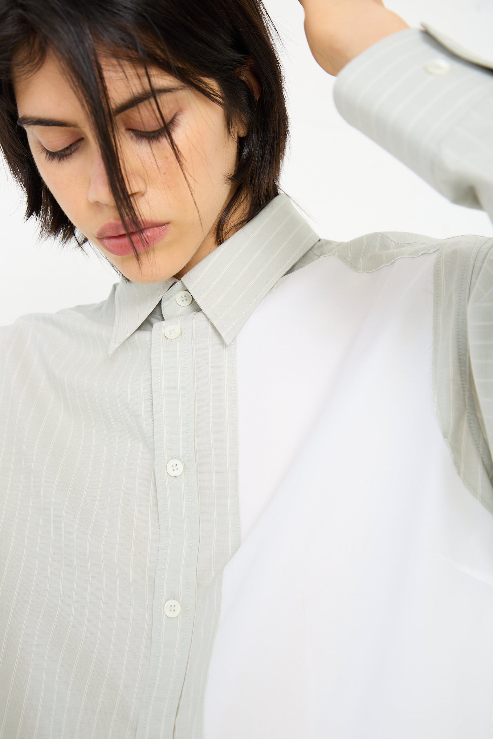 A person with short hair is wearing an MM6 Long Sleeved Shirt in Iguana, tilting their head slightly and looking downward. Their left arm is raised.