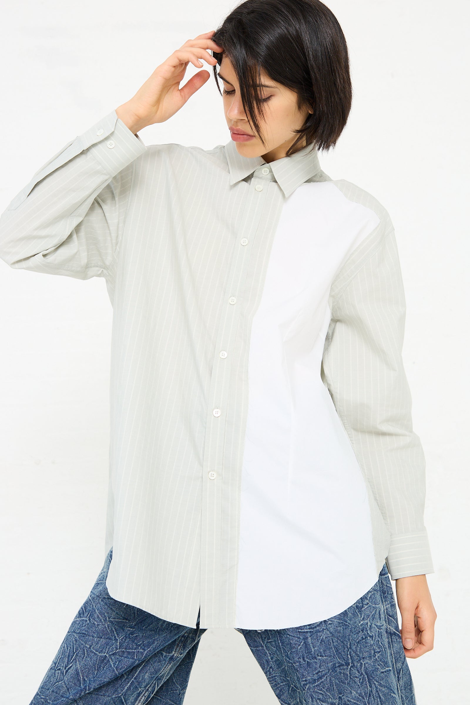 A person wearing a loose-fitting, MM6 Long Sleeved Shirt in Iguana and blue jeans poses with one hand on their head against a plain background.