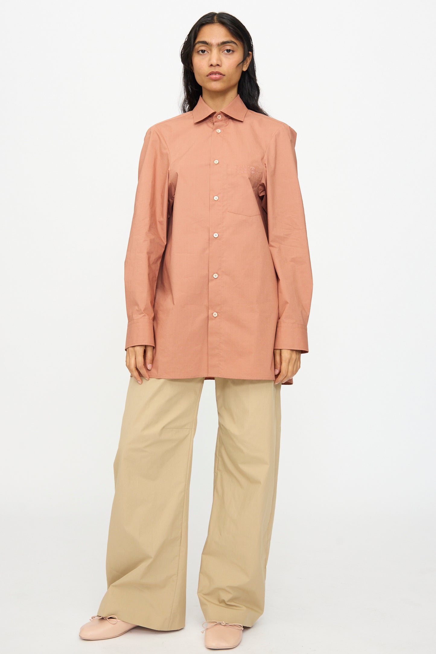 A person stands facing forward wearing the MM6 Long Sleeved Shirt in Terracotta Red, paired with wide-leg beige pants and light-colored shoes, set against a plain white background.