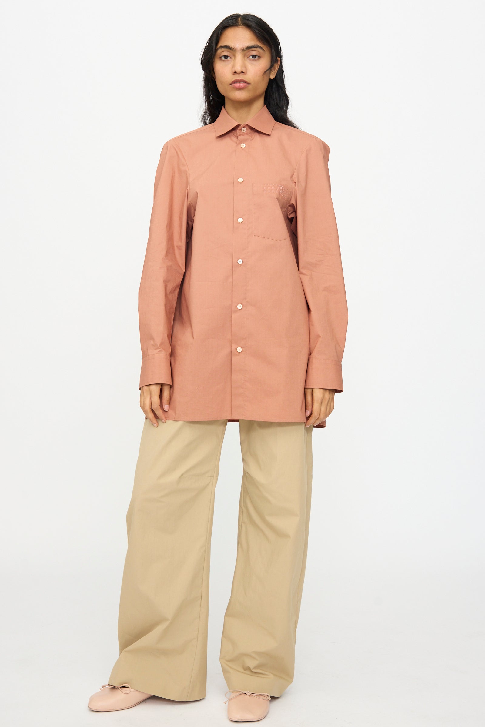 A person stands facing forward wearing the MM6 Long Sleeved Shirt in Terracotta Red, paired with wide-leg beige pants and light-colored shoes, set against a plain white background.