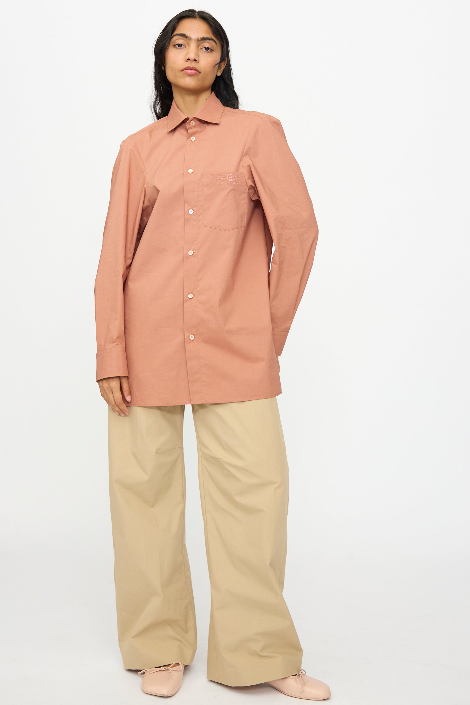 A person stands against a plain background wearing the MM6 Long Sleeved Shirt in Terracotta Red, paired with wide-leg beige pants and light pink shoes.