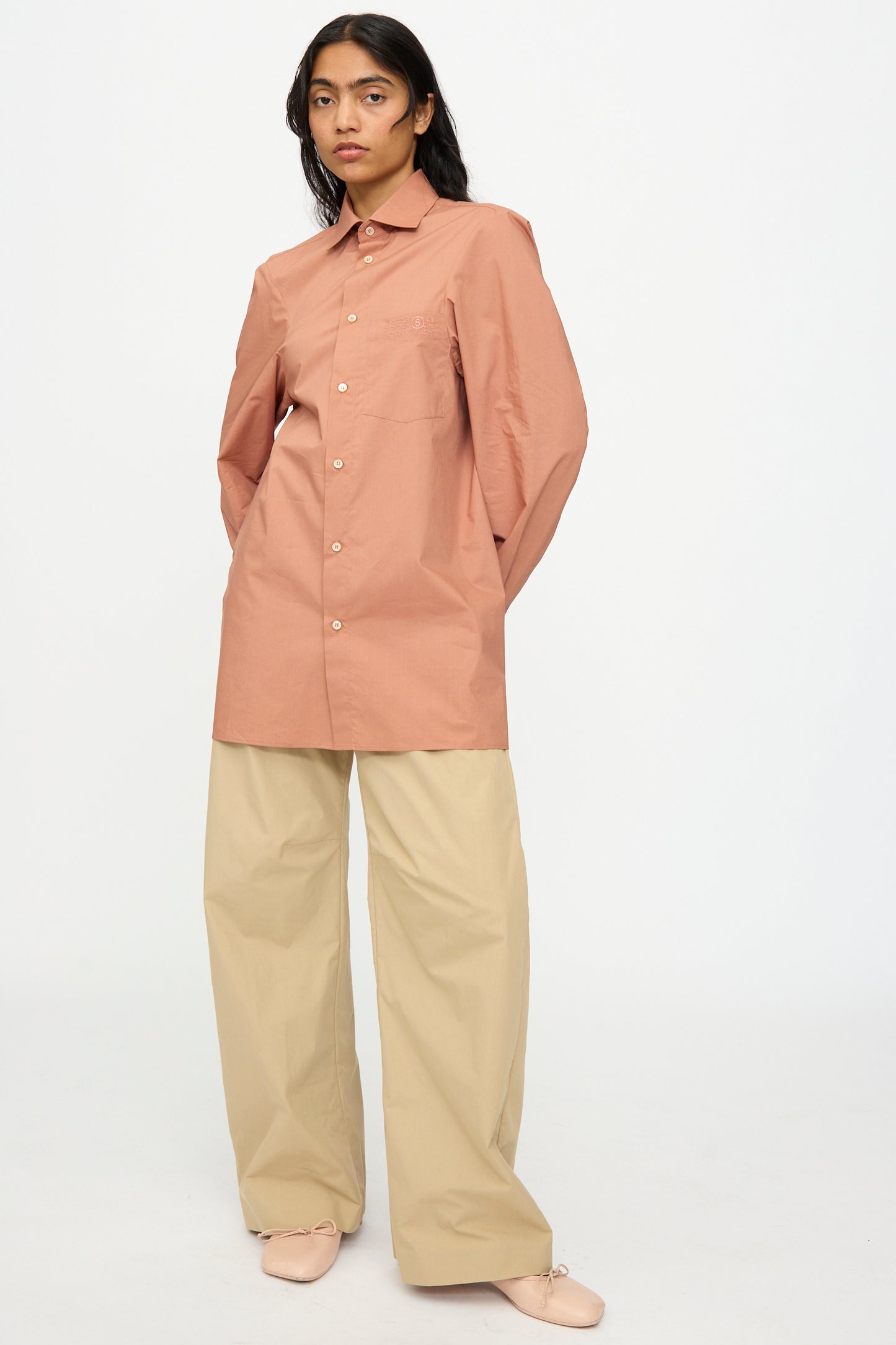 A person stands against a plain background wearing the MM6 Long Sleeved Shirt in Terracotta Red with loose beige pants and light-colored shoes.