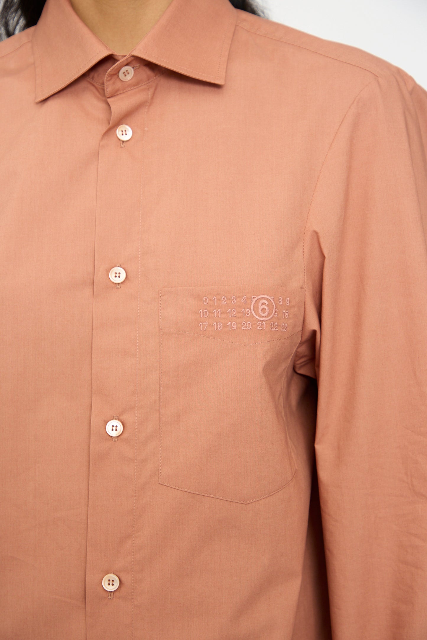 A person wears the MM6 Long Sleeved Shirt in Terracotta Red, featuring a relaxed fit, chest pocket, and small white numerical detail above the pocket.