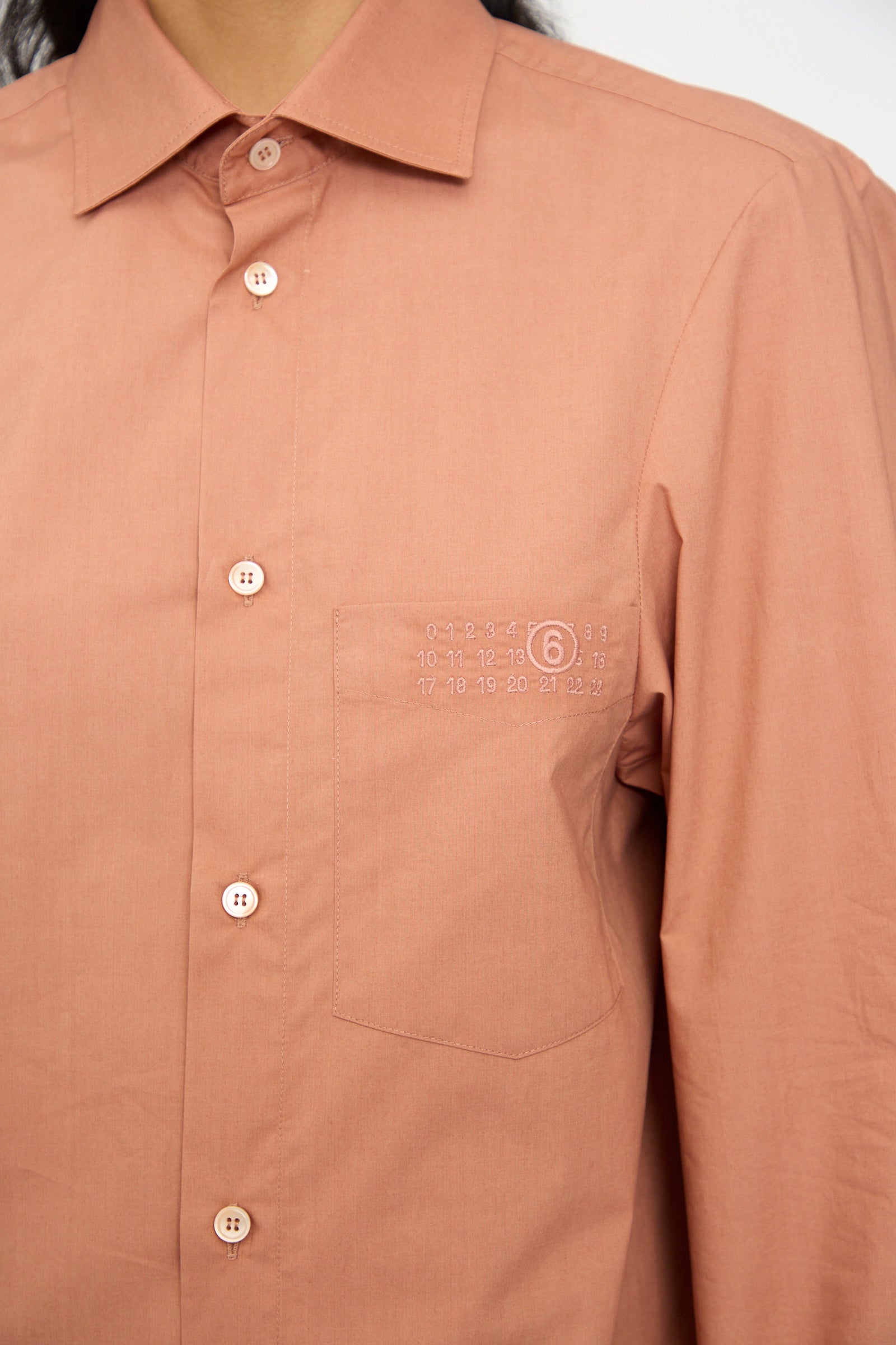 A person wears the MM6 Long Sleeved Shirt in Terracotta Red, featuring a relaxed fit, chest pocket, and small white numerical detail above the pocket.