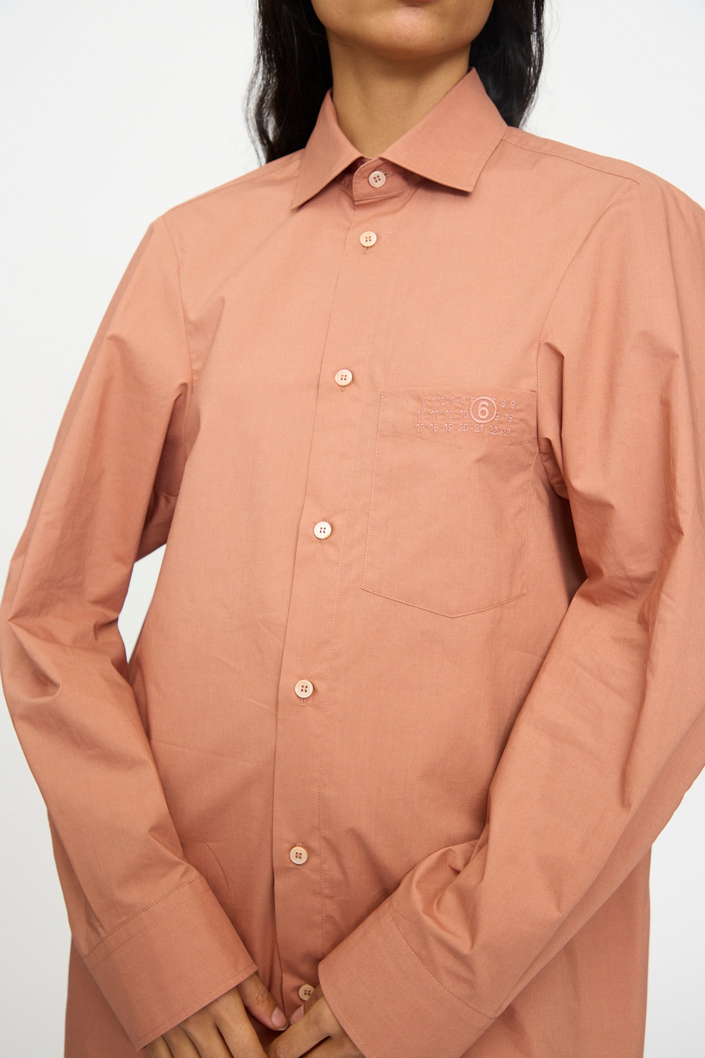 A person wears the MM6 Long Sleeved Shirt in Terracotta Red—relaxed fit with button-up front, chest pocket, and minimal embroidery—crafted from soft cotton poplin, standing against a plain background.