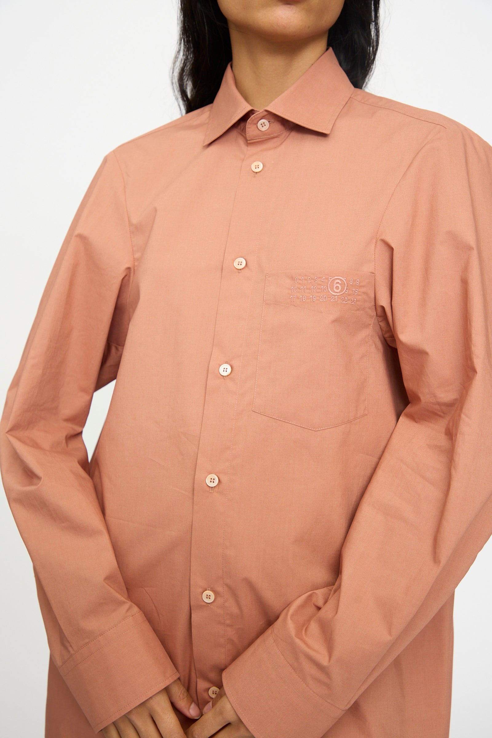 A person wears the MM6 Long Sleeved Shirt in Terracotta Red—relaxed fit with button-up front, chest pocket, and minimal embroidery—crafted from soft cotton poplin, standing against a plain background.