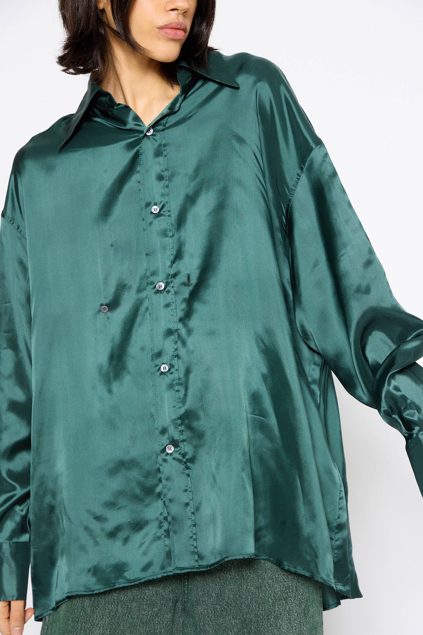 A person is wearing a Long Sleeved Shirt in Green by MM6, which features a shiny green satin fabric, a collar, and a relaxed fit. They are also wearing matching green pants.