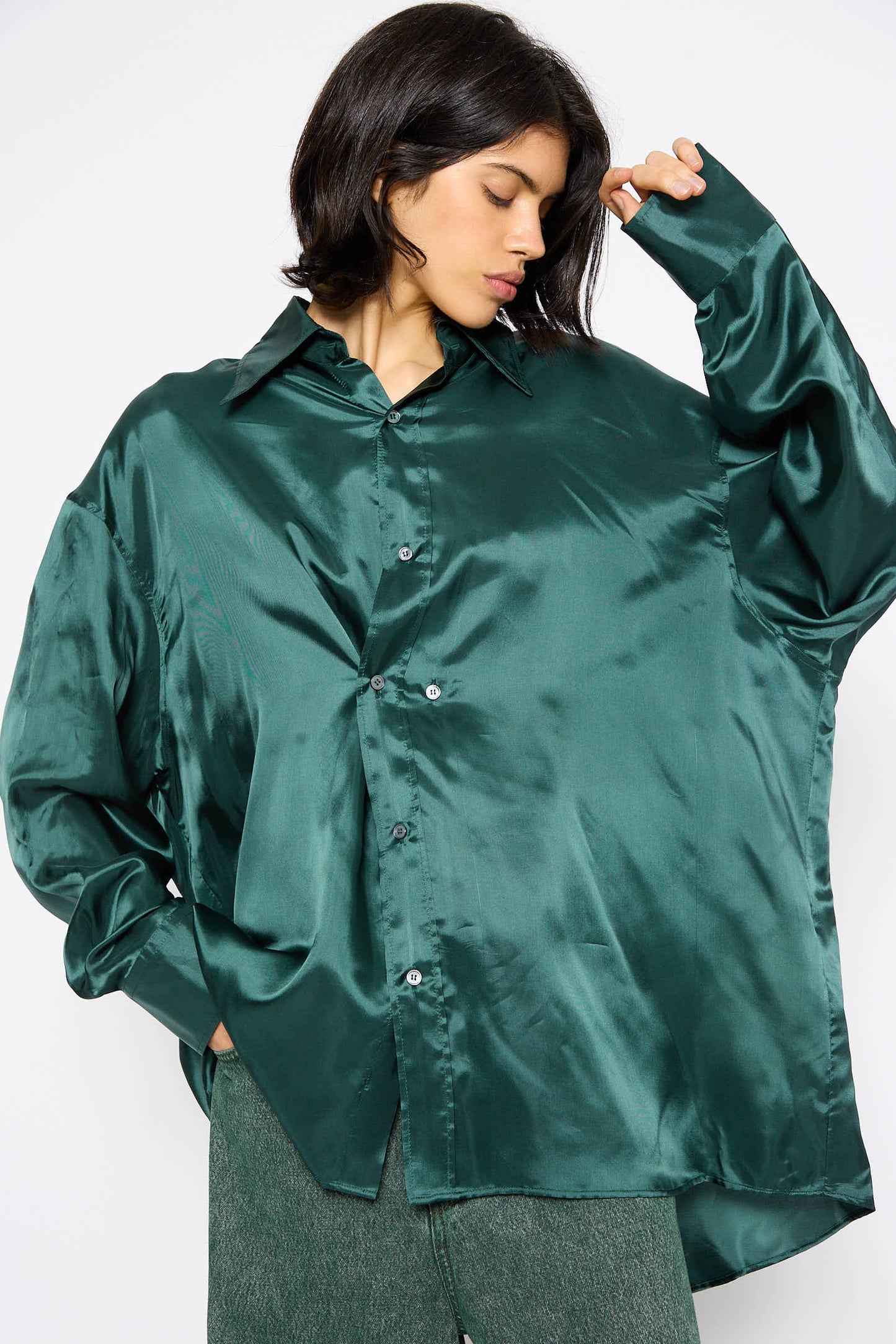 A person with dark hair wears an oversized, shiny green Long Sleeved Shirt in Green from MM6 and matching green pants, looking down with their hand resting on their forehead.