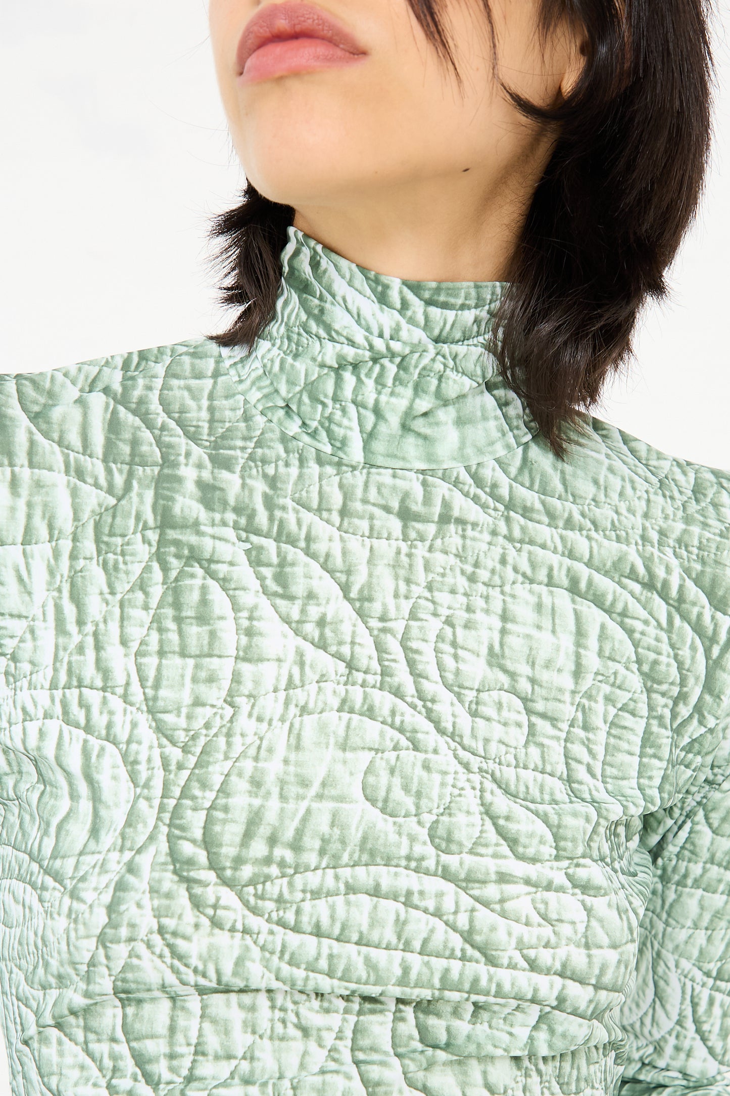 Close-up of a person wearing the MM6 Long Sleeved Top in Iguana, a fitted, light green turtleneck top with a high collar. The textured fabric features an iguana graphic print. The person's head is slightly tilted, showing their short, dark hair and part of the lower face.