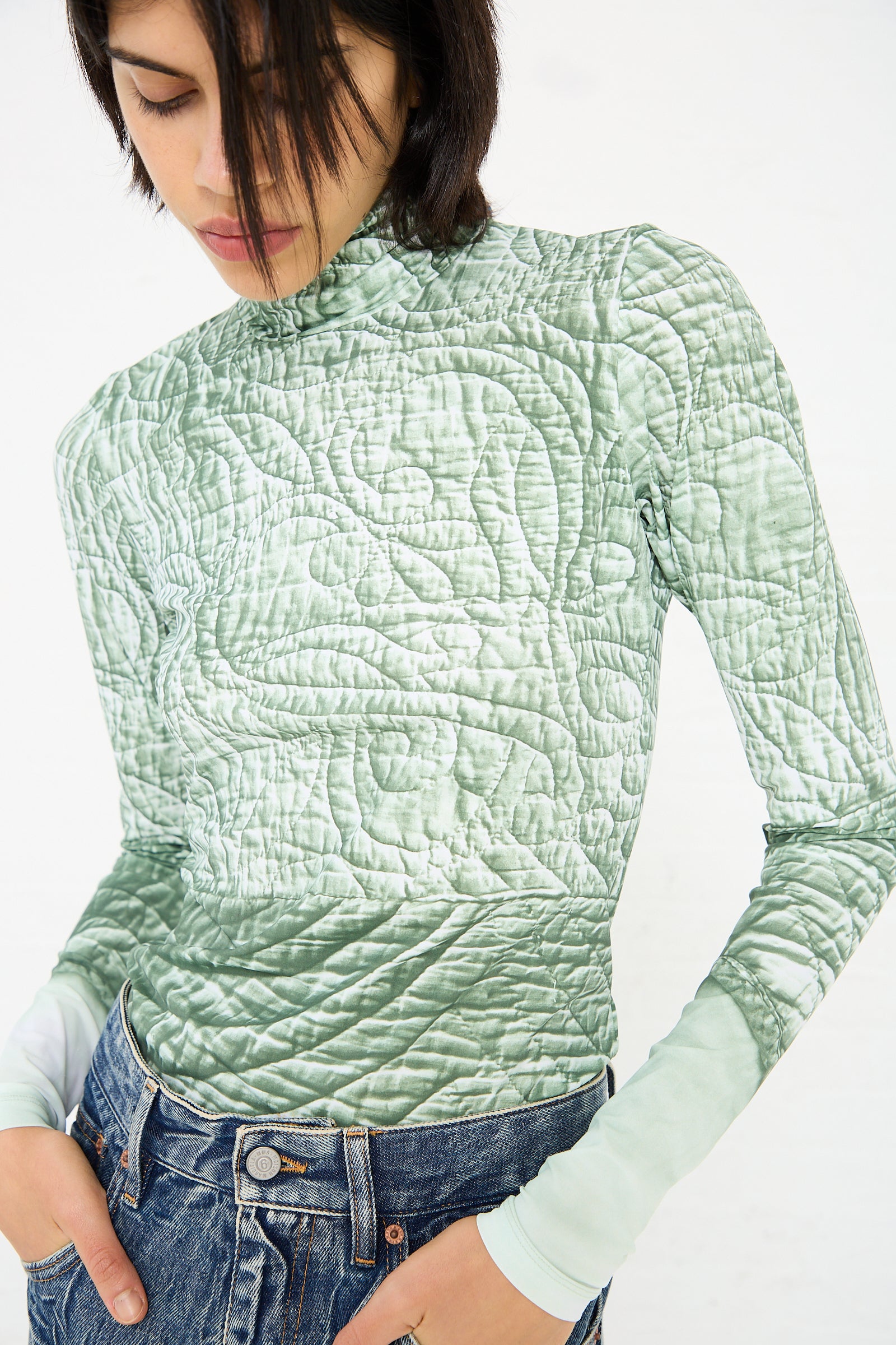 A person wearing an MM6 Long Sleeved Top in Iguana and blue jeans is looking down, with chin-length dark hair partially covering their face.