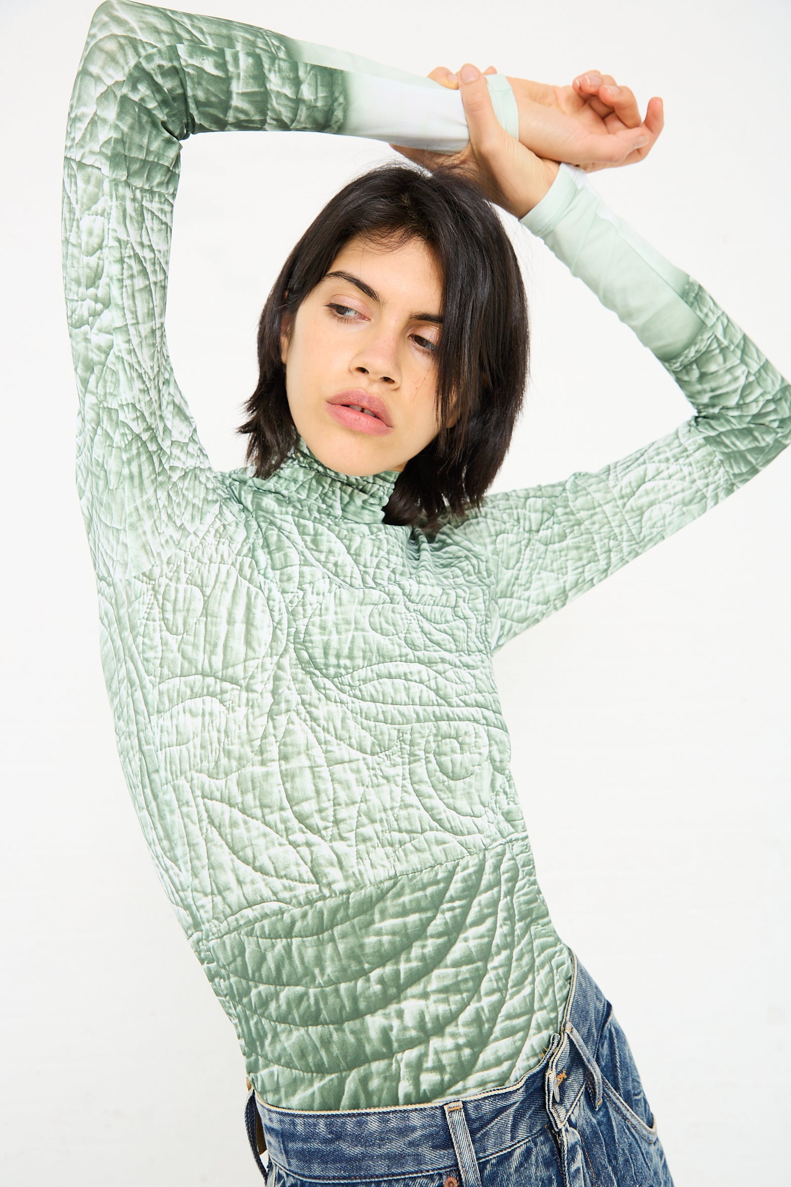 A person with black hair in a green, iguana-print top stretches with their arms above their head. They are wearing the Long Sleeved Top in Iguana by MM6, blue jeans, and standing against a plain white background.