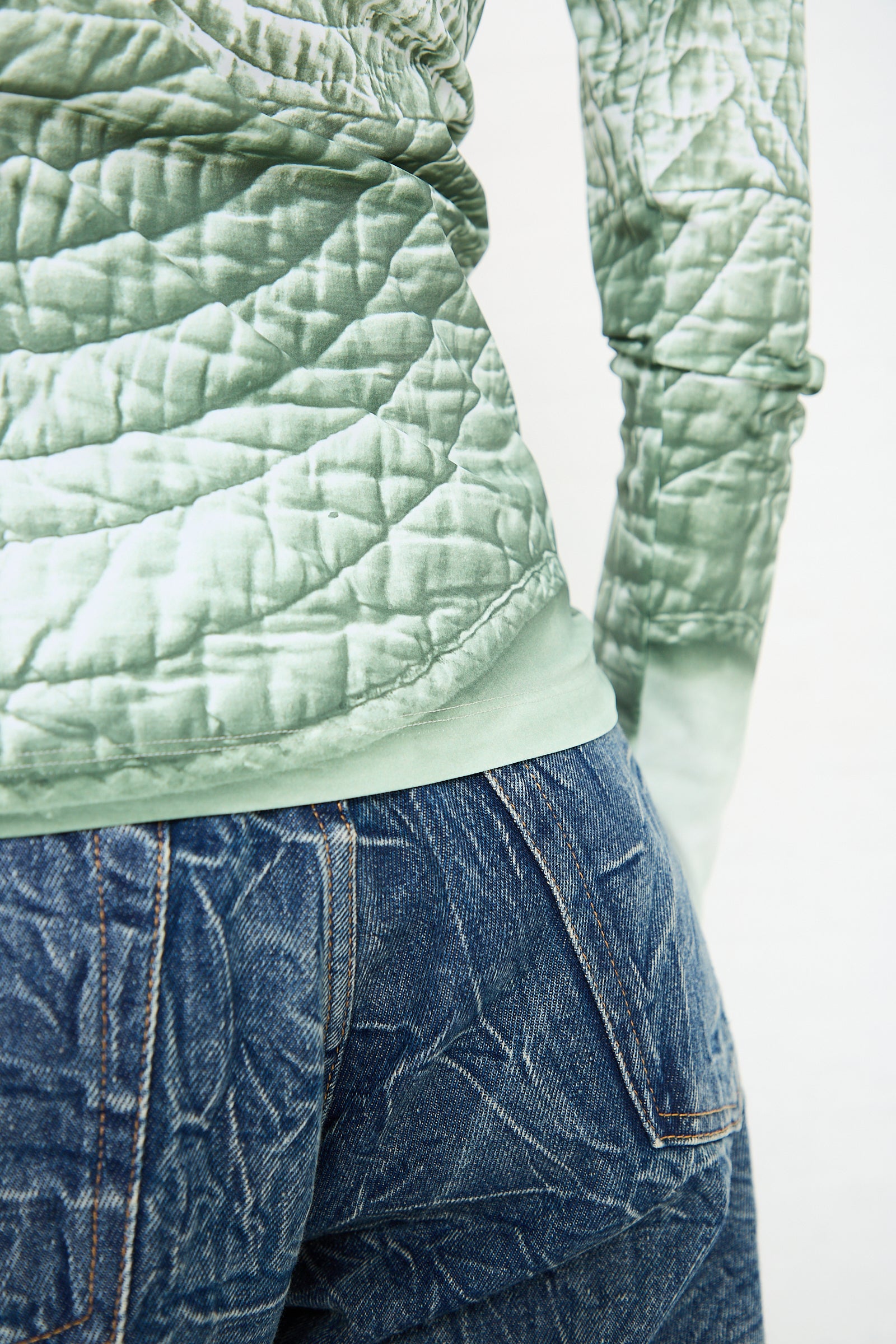 Close-up of a person wearing a green, long sleeved top in iguana by MM6 tucked into blue, textured jeans.