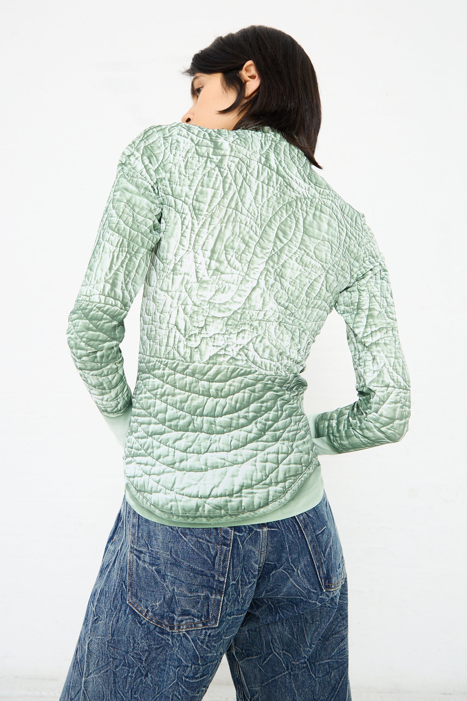 A person with short dark hair is facing away, wearing a MM6 Long Sleeved Top in Iguana and blue jeans against a plain white background.